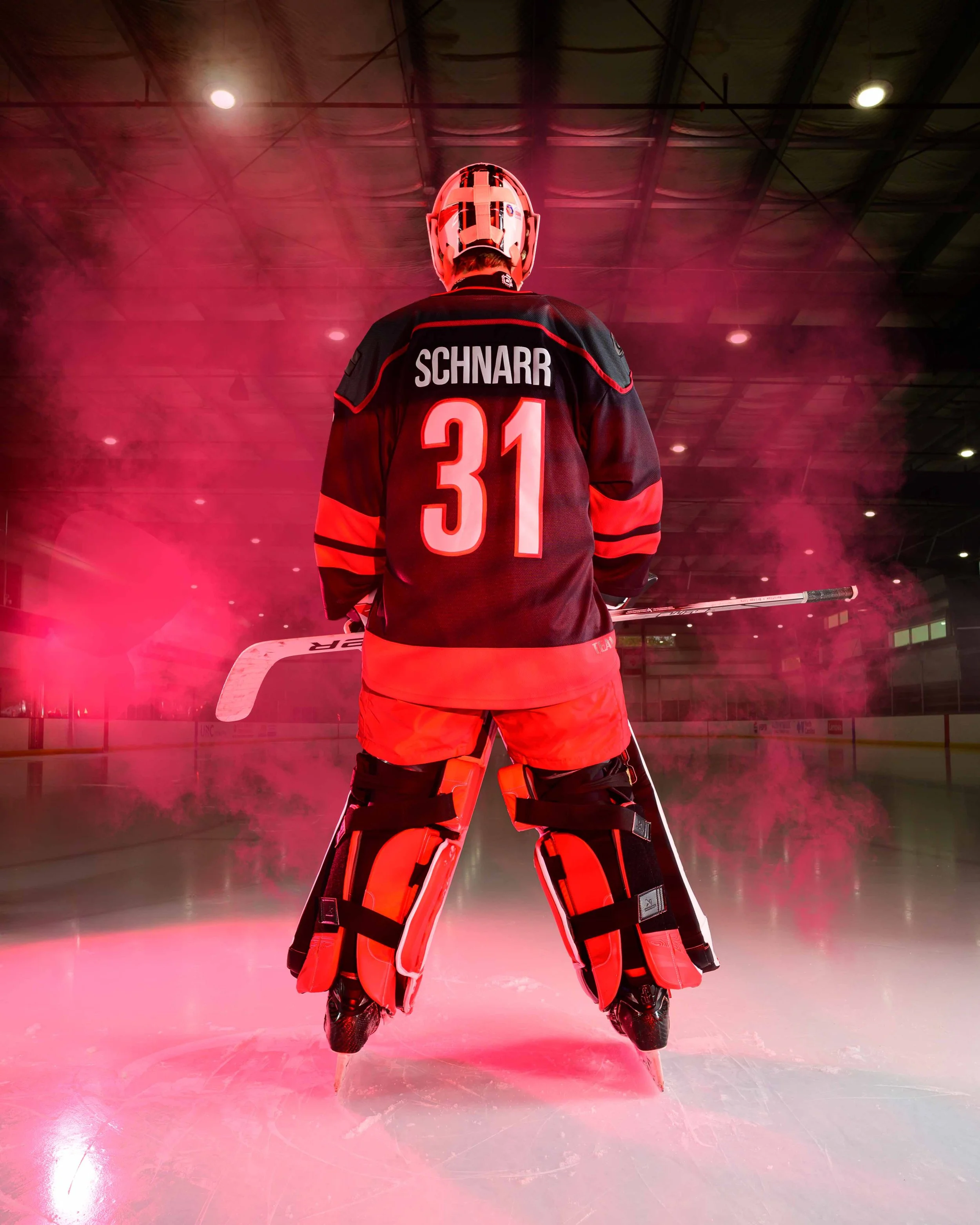 Ice hockey goalie standing on the ice rink with their back facing the camera. The goalie is wearing a black and red uniform with the number 31 and the name 'SCHNARR' on the back. The scene is illuminated with red lighting, creating a smoky atmosphere