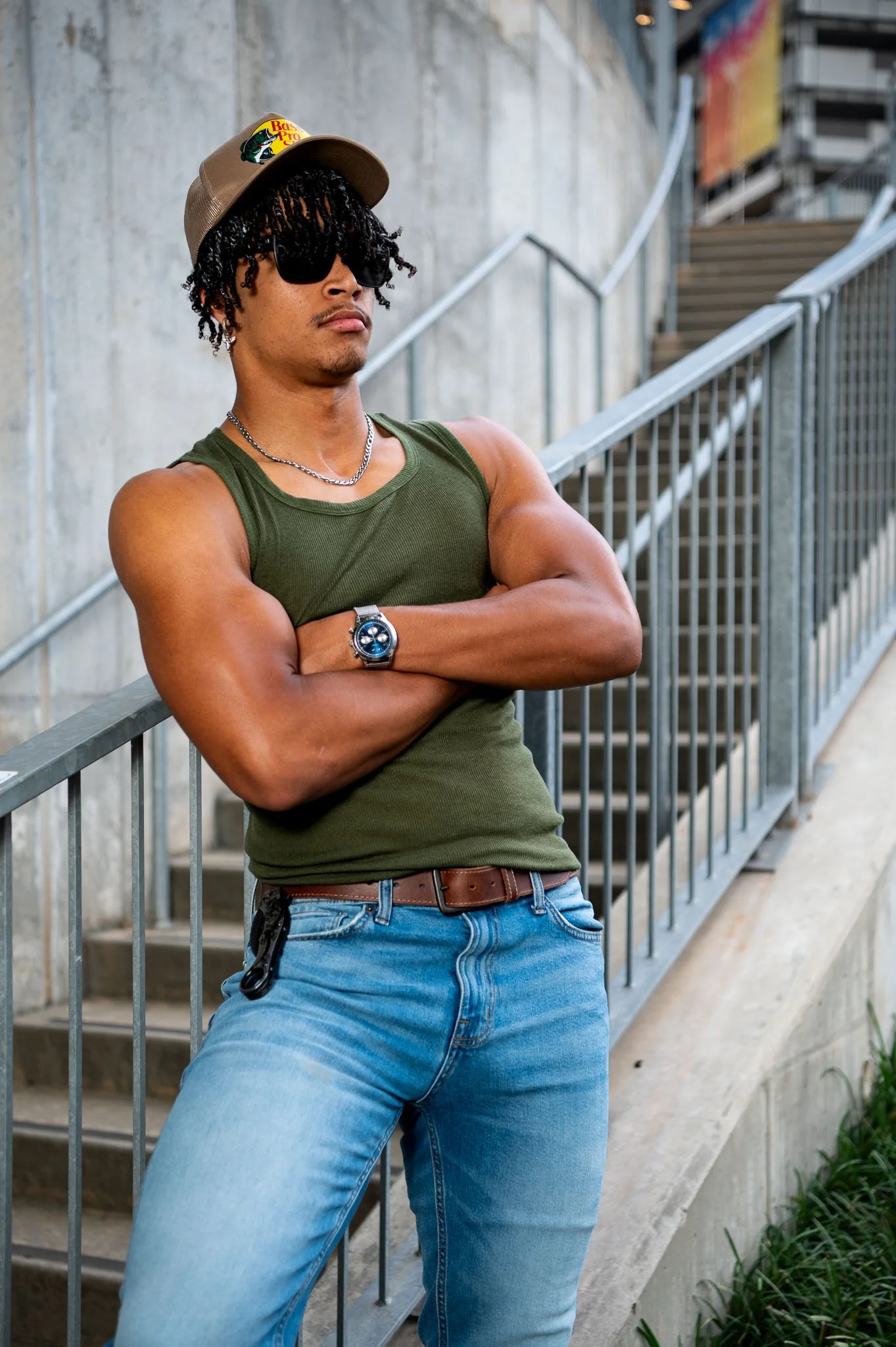 Young man with dreadlocks, wearing sunglasses, a cap with a Bass Pro Shops logo, an olive green tank top, jeans, and a wristwatch, standing with crossed arms against a metal railing outside.