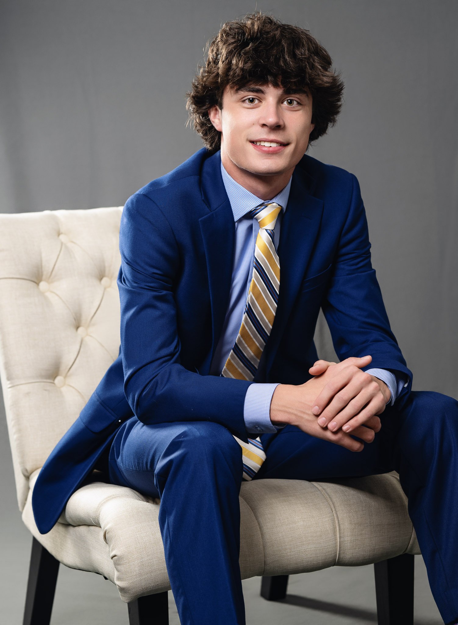 A young man with curly brown hair in a blue suit, shirt, and yellow striped tie sitting on a beige chair against a gray background.