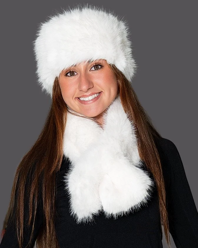 Woman smiling wearing a white furry winter hat and matching scarf against a gray background.
