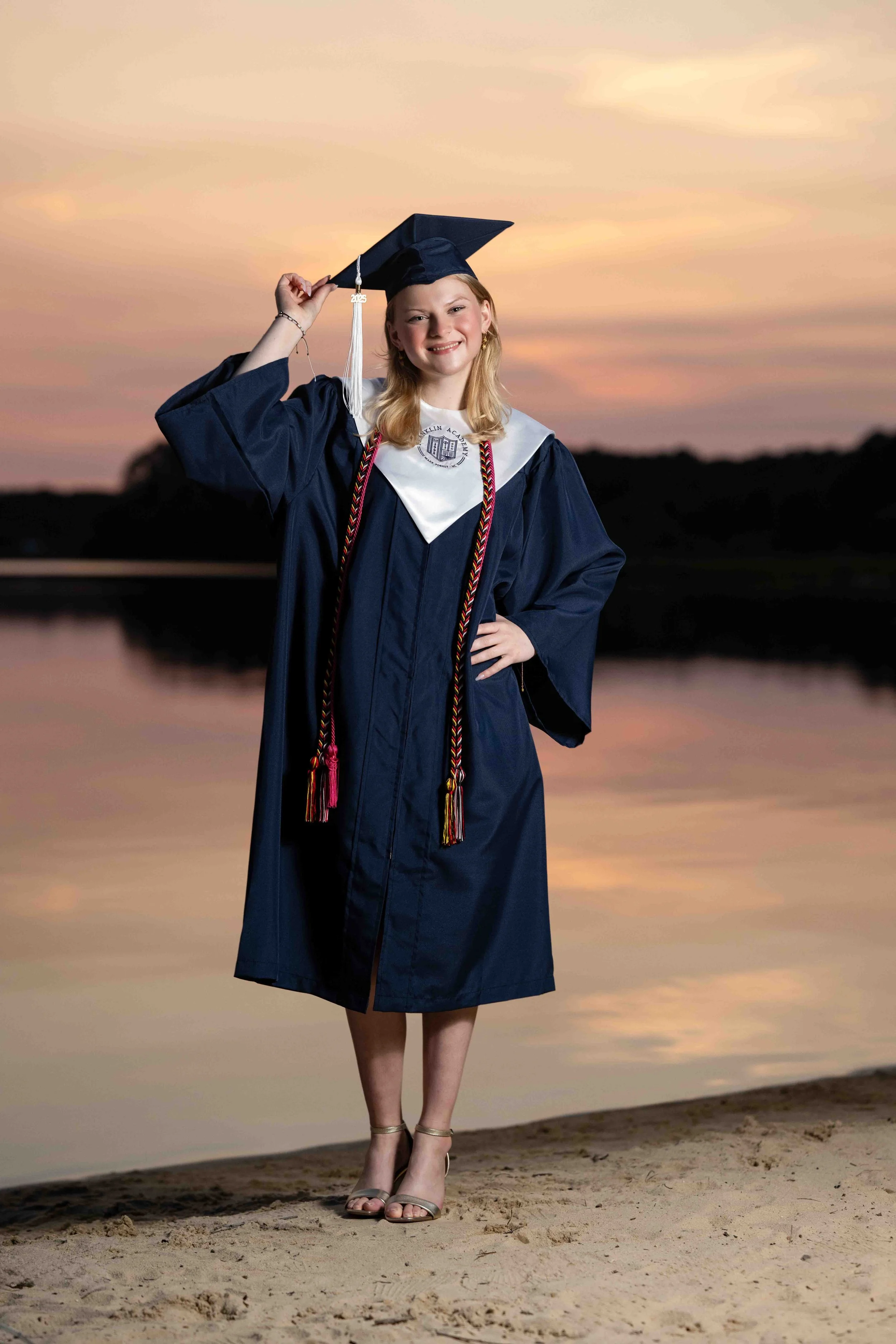 A young woman in graduation gown and cap standing on a sandy beach at sunset, smiling and posing with one hand on her hip and the other adjusting her cap, with a calm body of water in the background.