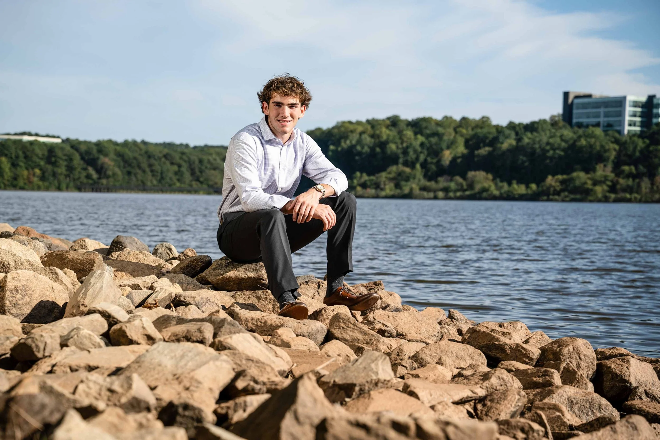 A young man in business attire sitting on rocks by a lake during daytime.