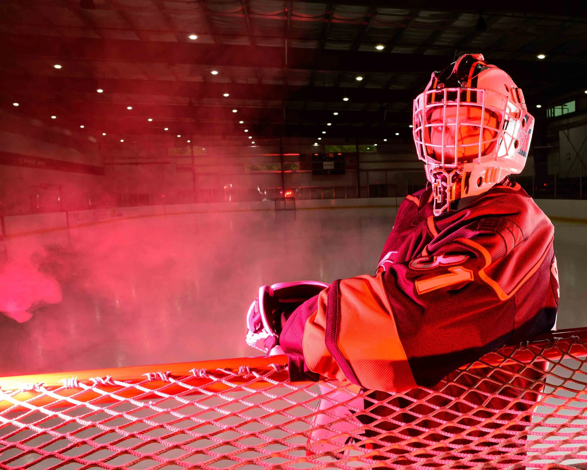 A hockey goalie in full gear, sitting on the bench at an indoor ice rink with red lighting and pink hockey smoke.