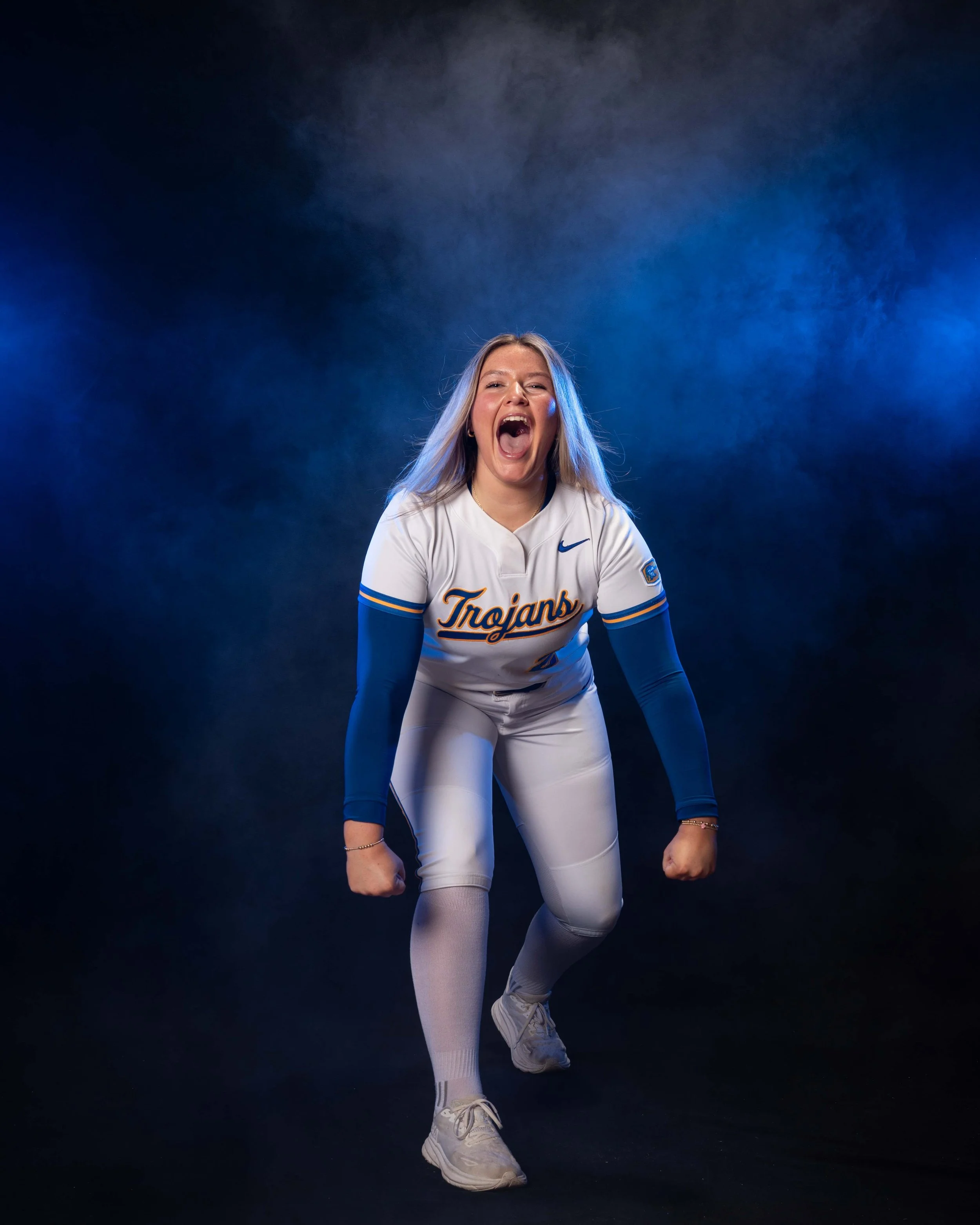Young woman in a football uniform with 'Trojans' on the jersey, celebrating with clenched fists and an open mouth, against a dark, smoky background.