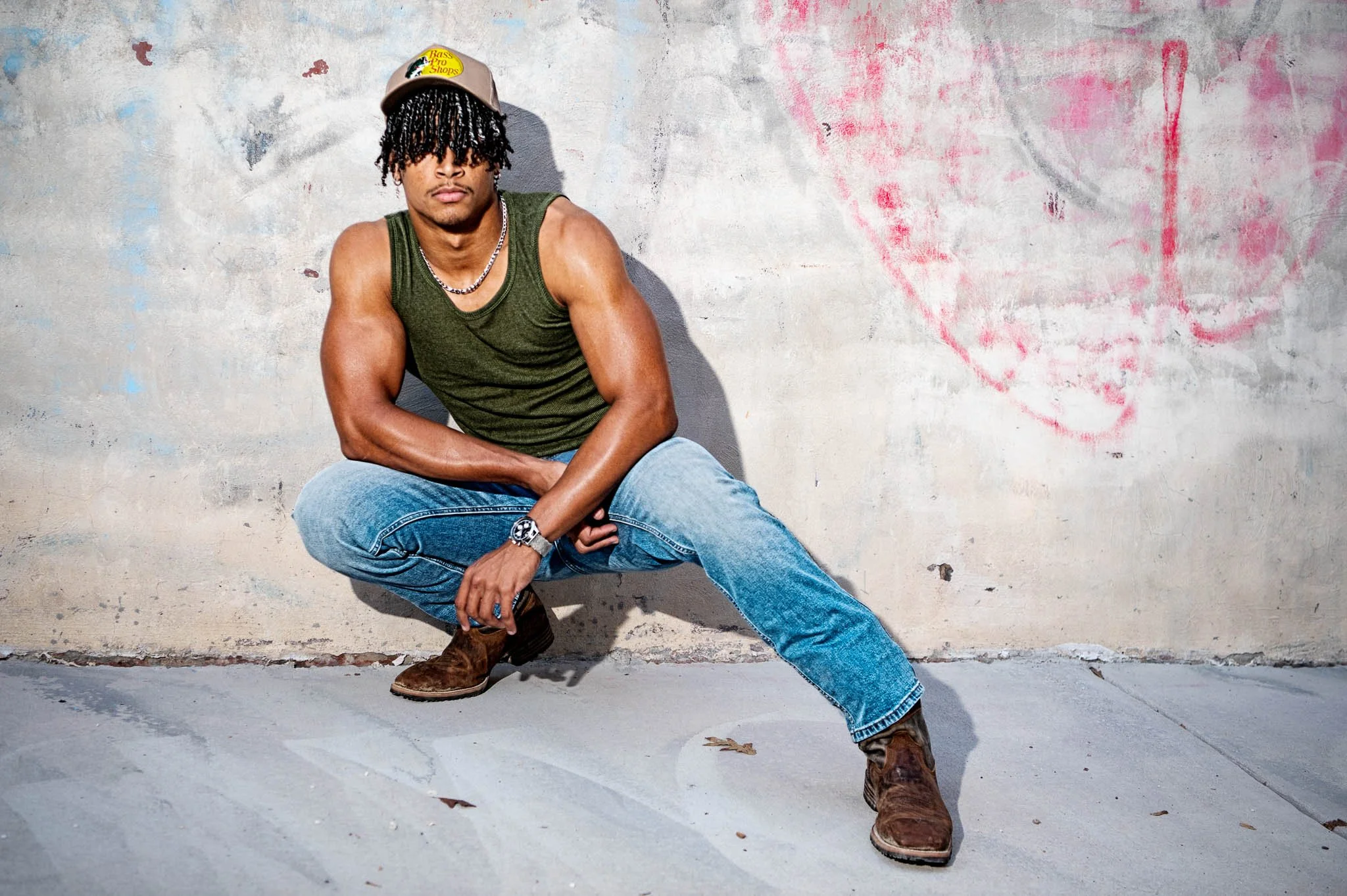 A man with dreadlocks wearing a beige cap, green tank top, blue jeans, and brown boots, squatting against a weathered wall with graffiti.