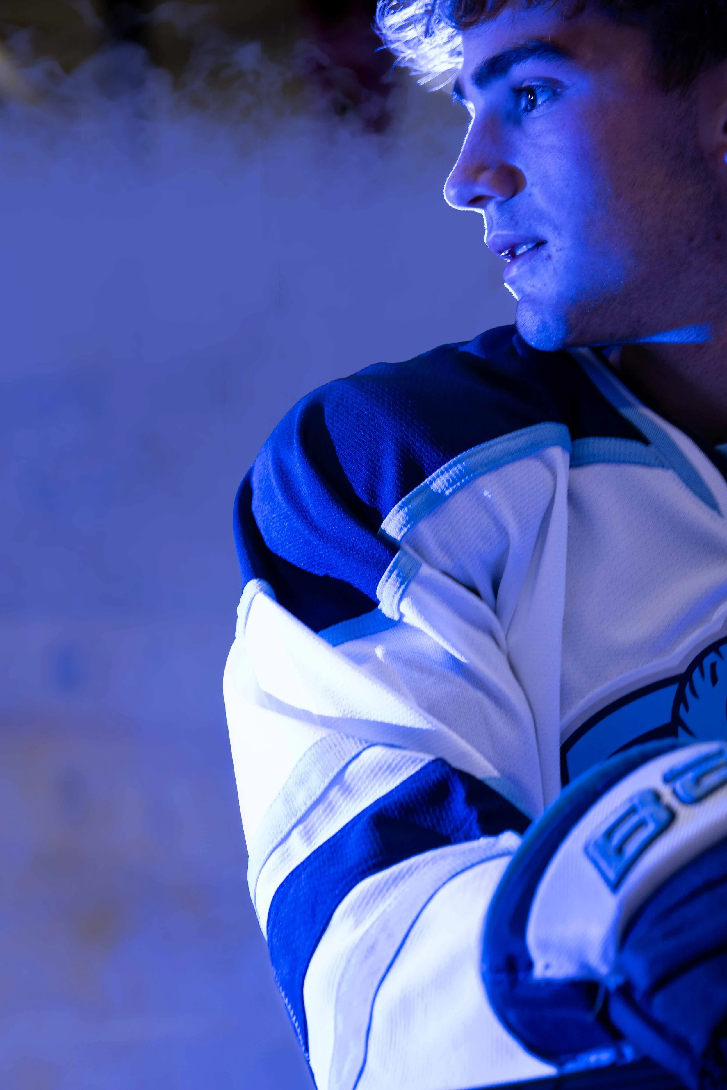 Close-up of a young male hockey player in a white and blue jersey, with blue lighting and smoke in the background.