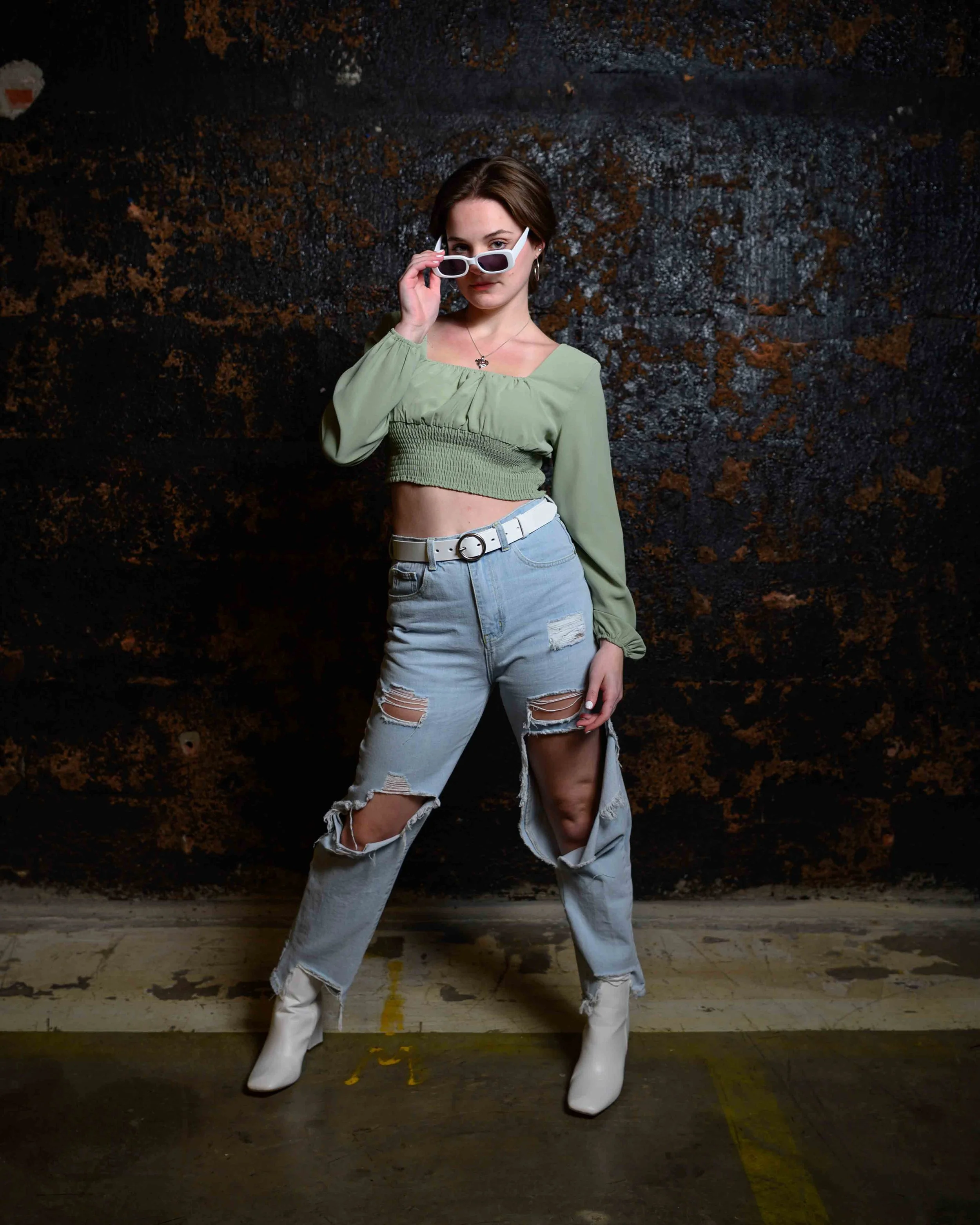 A young woman standing in front of a dark, textured background, wearing a green crop top, ripped light blue jeans, white cowboy boots, and white sunglasses, with a confident pose and expression.