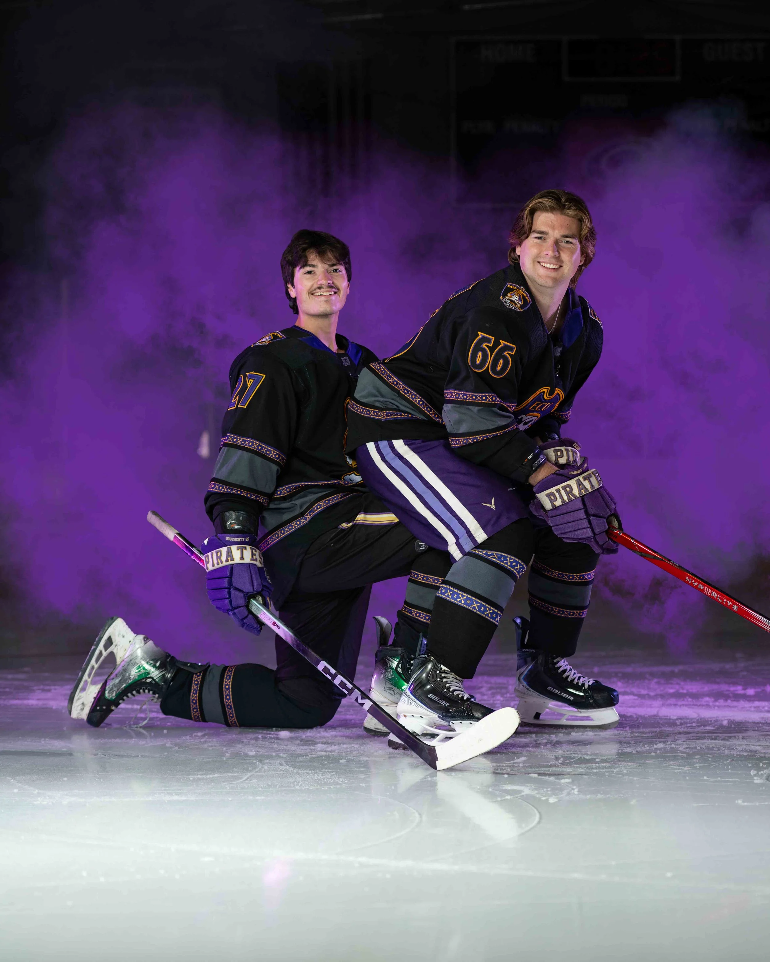 Two ice hockey players in black jerseys with purple and gold accents, kneeling on ice rink, smiling, with purple fog effects in the background.