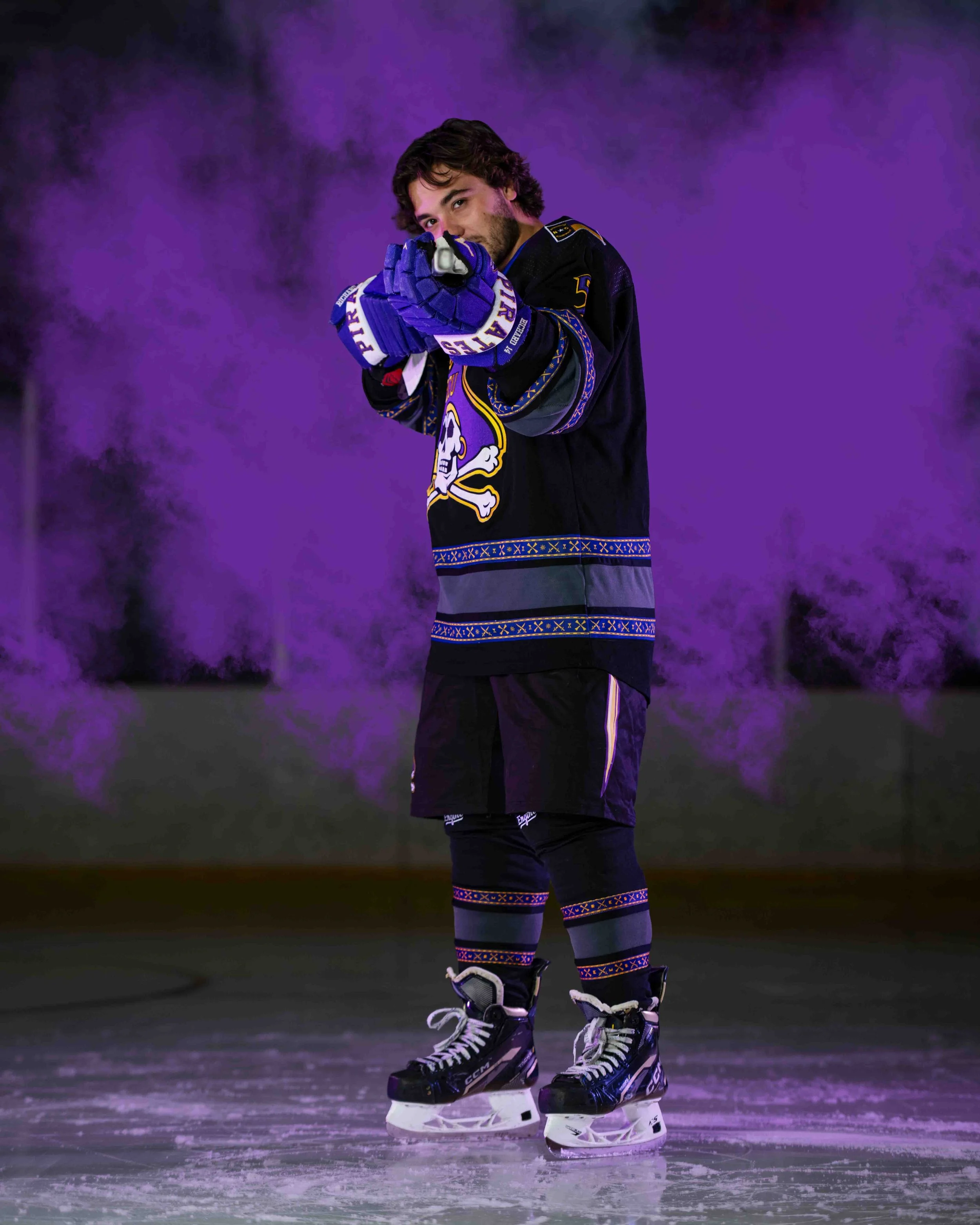 A man wearing hockey gear, including skates, gloves, and a jersey, is kneeling on the ice rink pointing a hockey stick towards the camera. The background features purple mist or smoke.