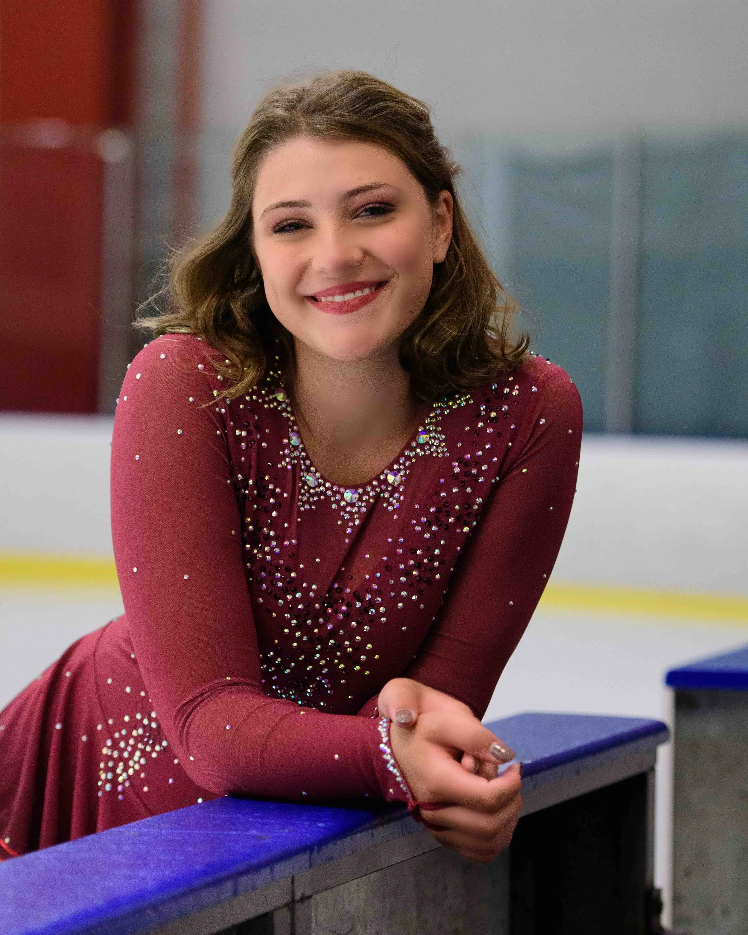 A young woman in a red figure skating dress decorated with rhinestones, smiling, leaning on the barrier at an ice rink.