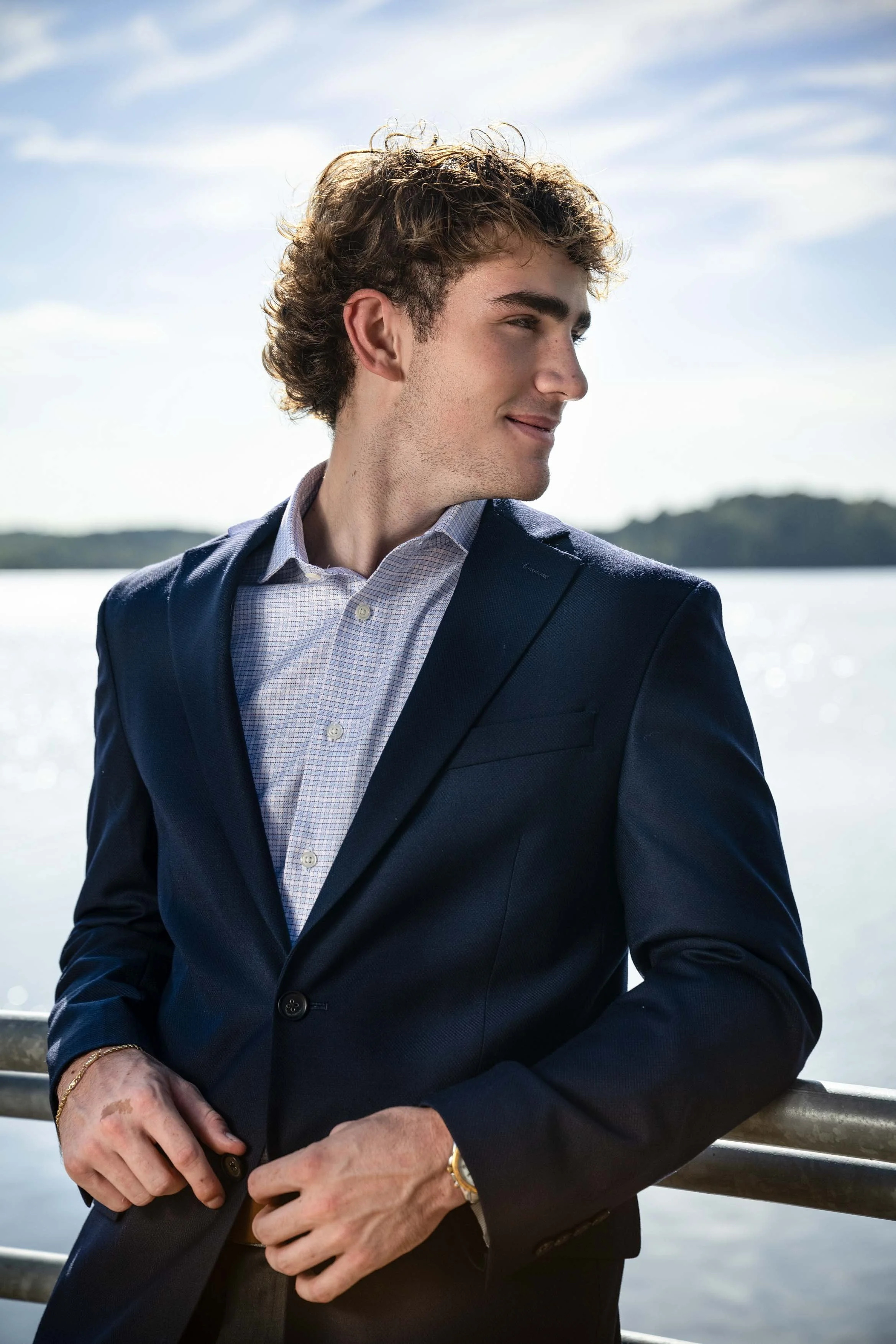 A young man in a navy blue suit and light checkered shirt standing outdoors near a body of water, with a scenic background of water and distant land, looking off to the side with a slight smile.