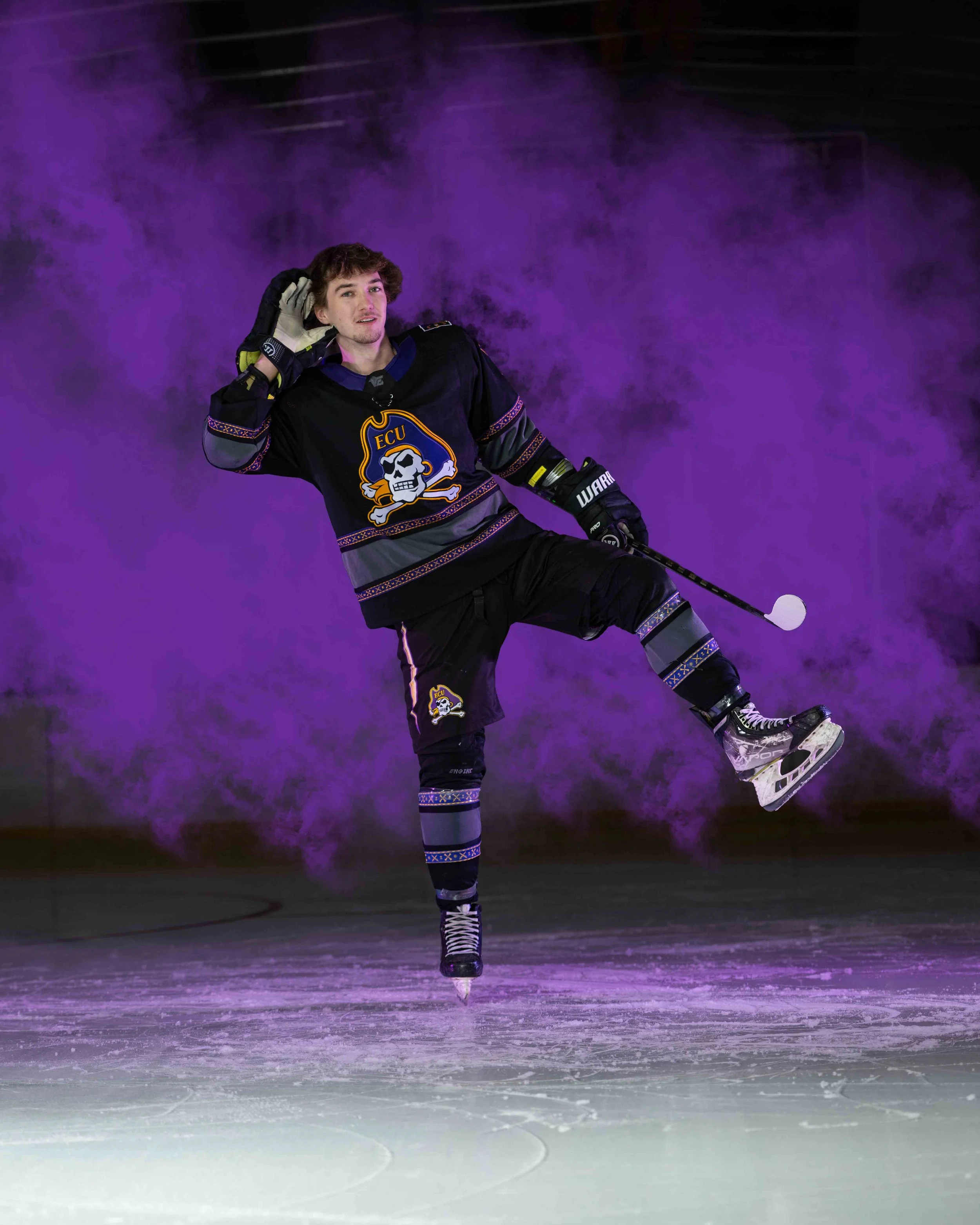 A young man wearing a black hockey jersey with an ECU skull logo, black hockey gloves, and ice skates is skating on an ice rink. He is performing a figure skating move and has one leg extended behind him. Purple fog or lighting surrounds him on the i