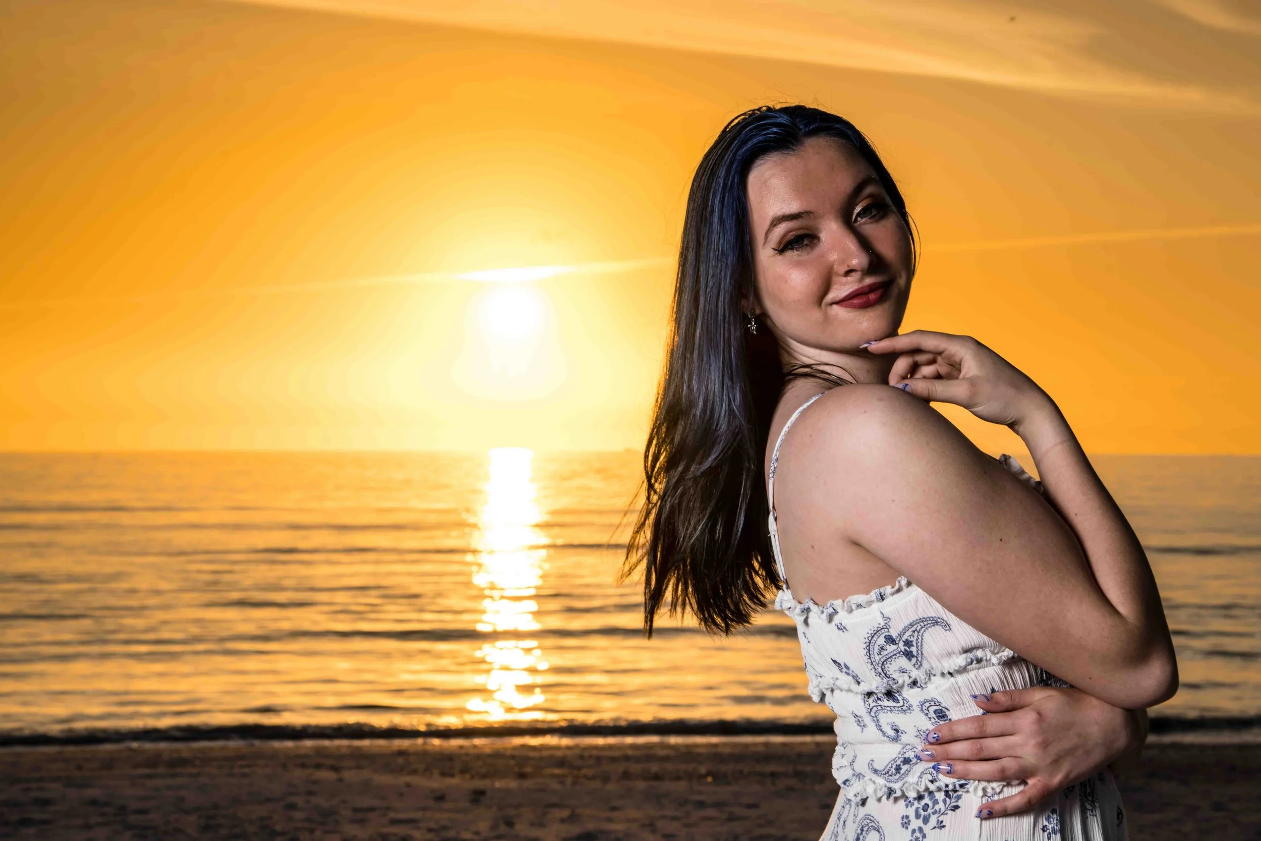 A woman with dark hair, wearing a white dress with blue patterns, standing on a beach during sunset, looking at the camera with a slight smile, with the sun setting over the ocean in the background.
