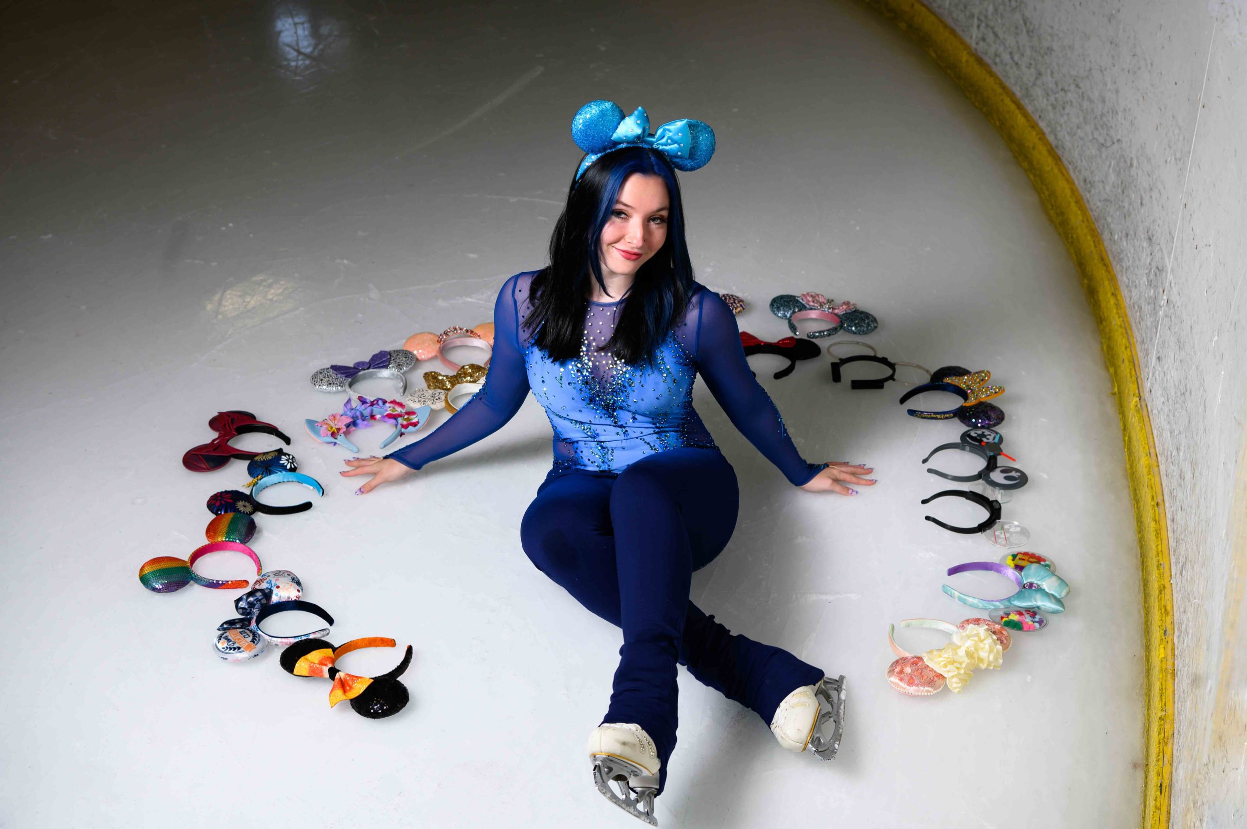 A woman with long black and blue hair, wearing a blue sequined top, black pants, white ice skates, and a blue Mickey Mouse ears headband, sitting on the ice surrounded by colorful headbands arranged in a circle.