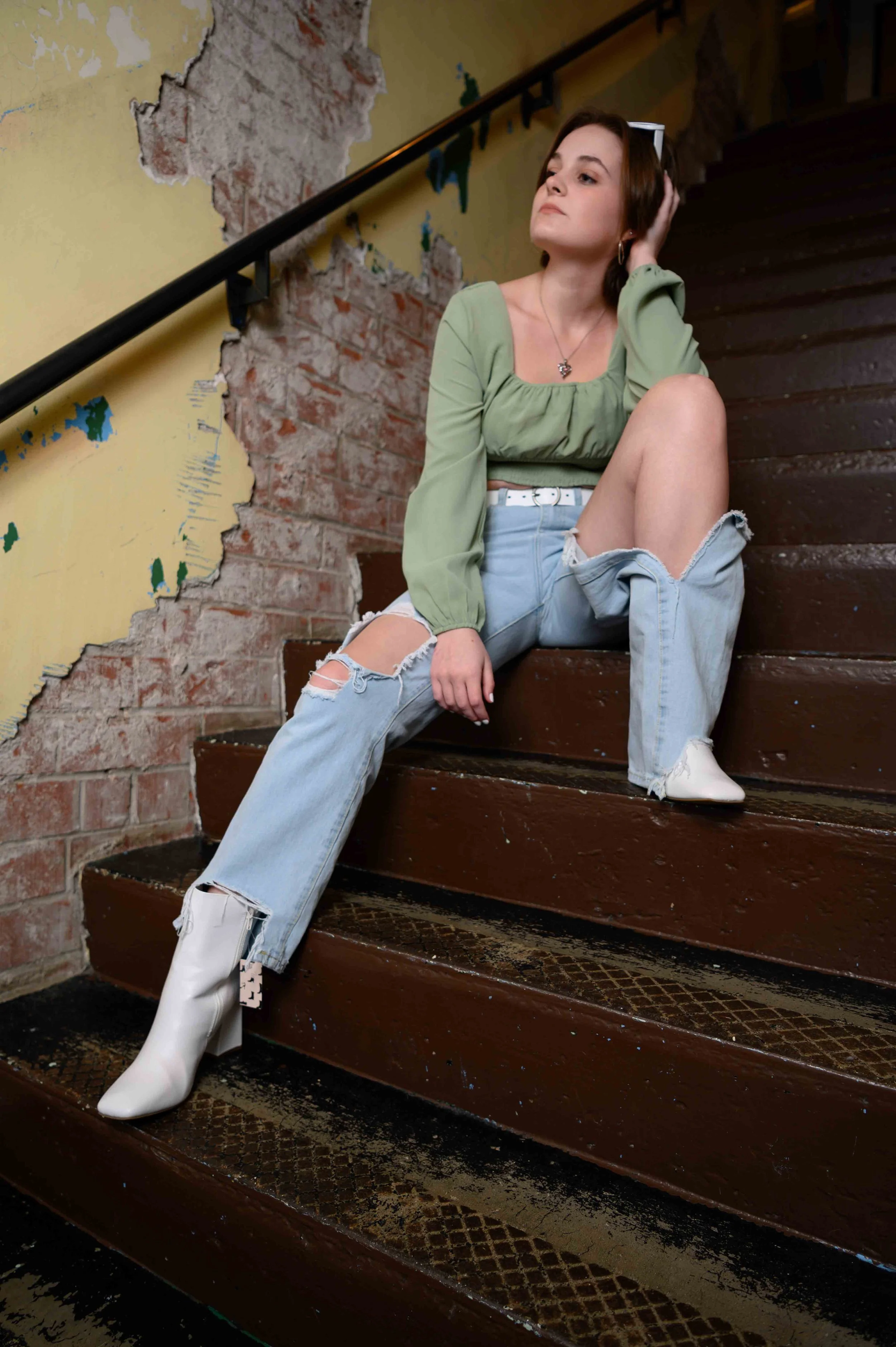 A young woman sitting on stairs inside a building with peeling yellow paint and exposed brick wall, wearing a green long-sleeve top, ripped jeans, and white boots.