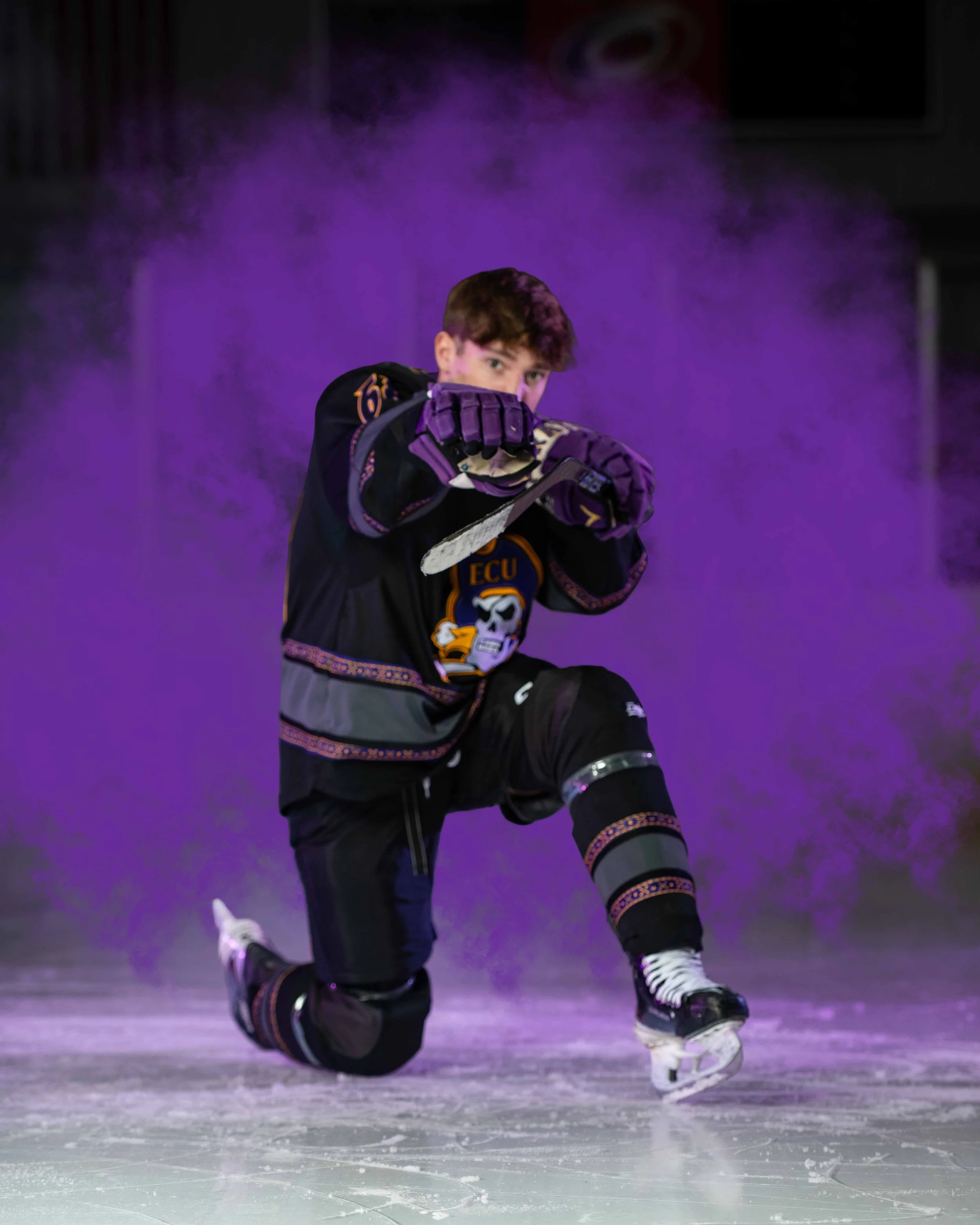 A young male figure skater kneeling on ice, wearing sports gear with purple, black, and orange colors and a skull logo. The background shows a purple-lit, smoky atmosphere.
