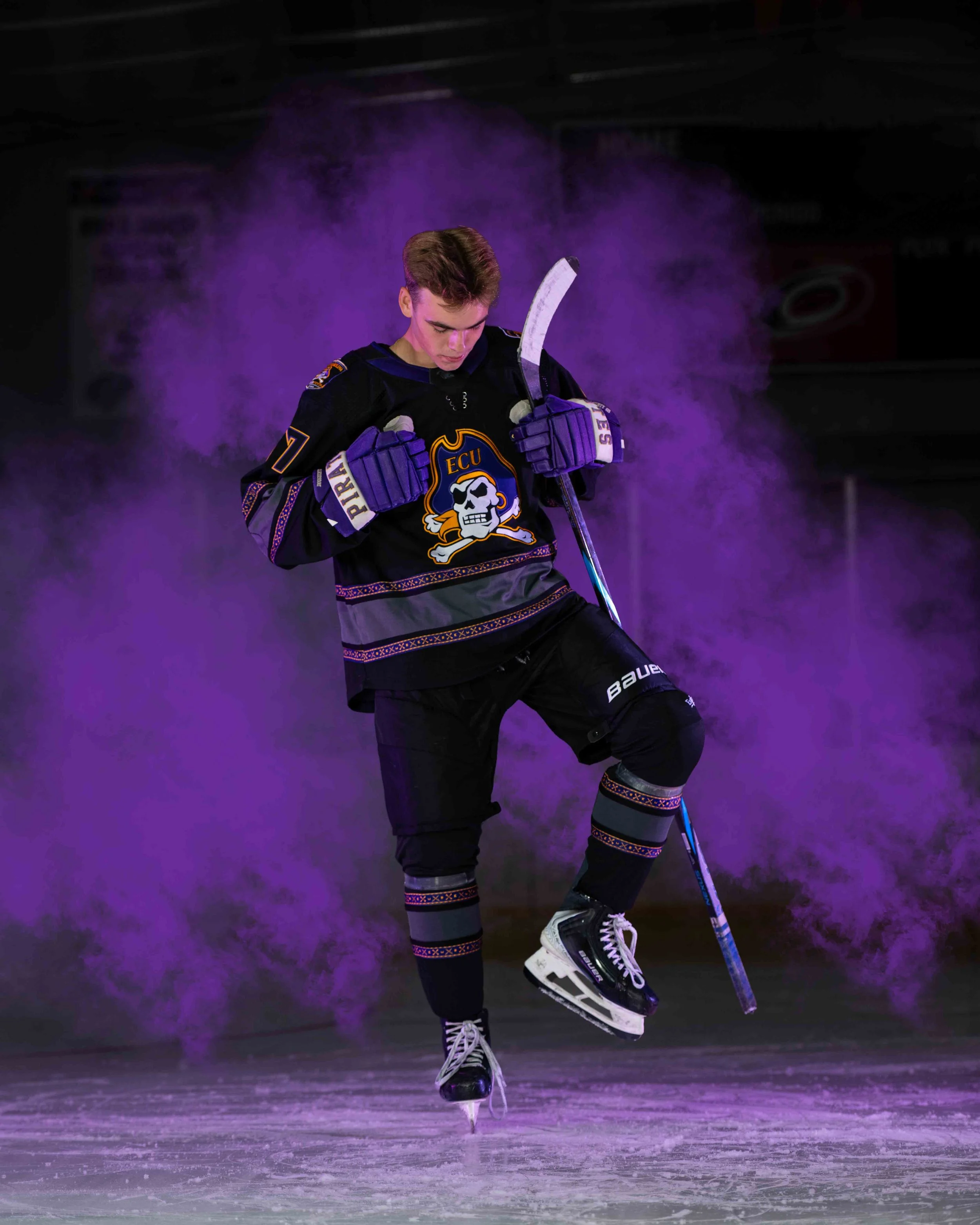 A young male ice hockey player in black and purple uniform performing a skating move on the ice, surrounded by purple smoke.