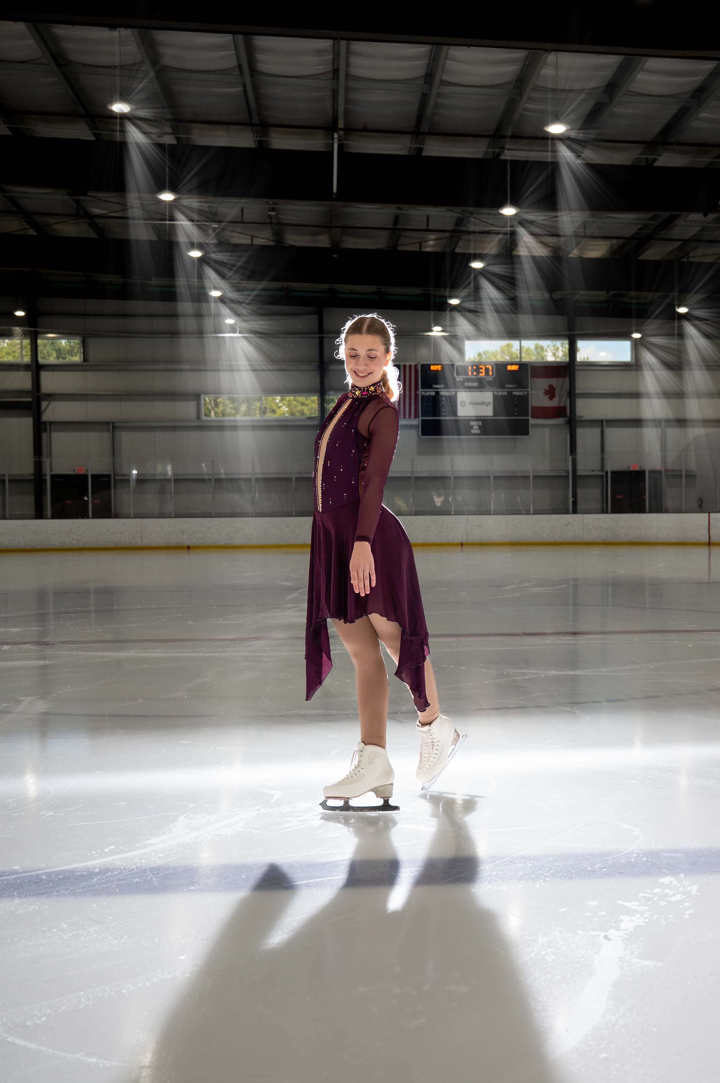 A young female figure skater in a purple dress performs on an ice rink with artificial beams of light overhead and a digital scoreboard in the background.