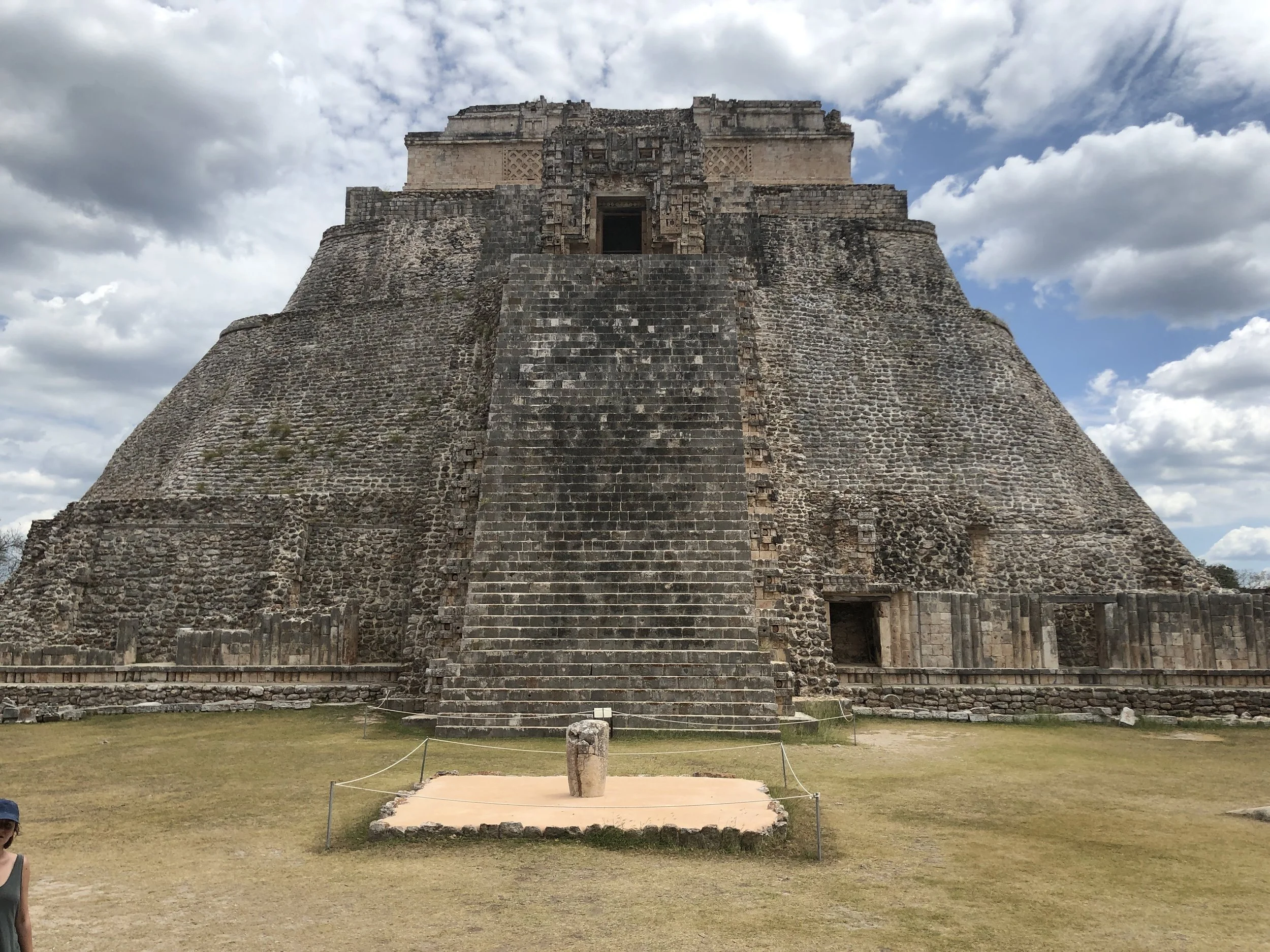 Ancient Mayan pyramid with stairs leading to the top, set against a blue sky with clouds, surrounded by grass.