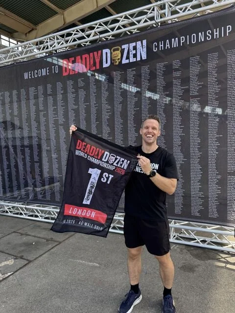 A smiling man holds a 1st place flag in front of a Deadly Dozen Championship event banner listing athlete names.