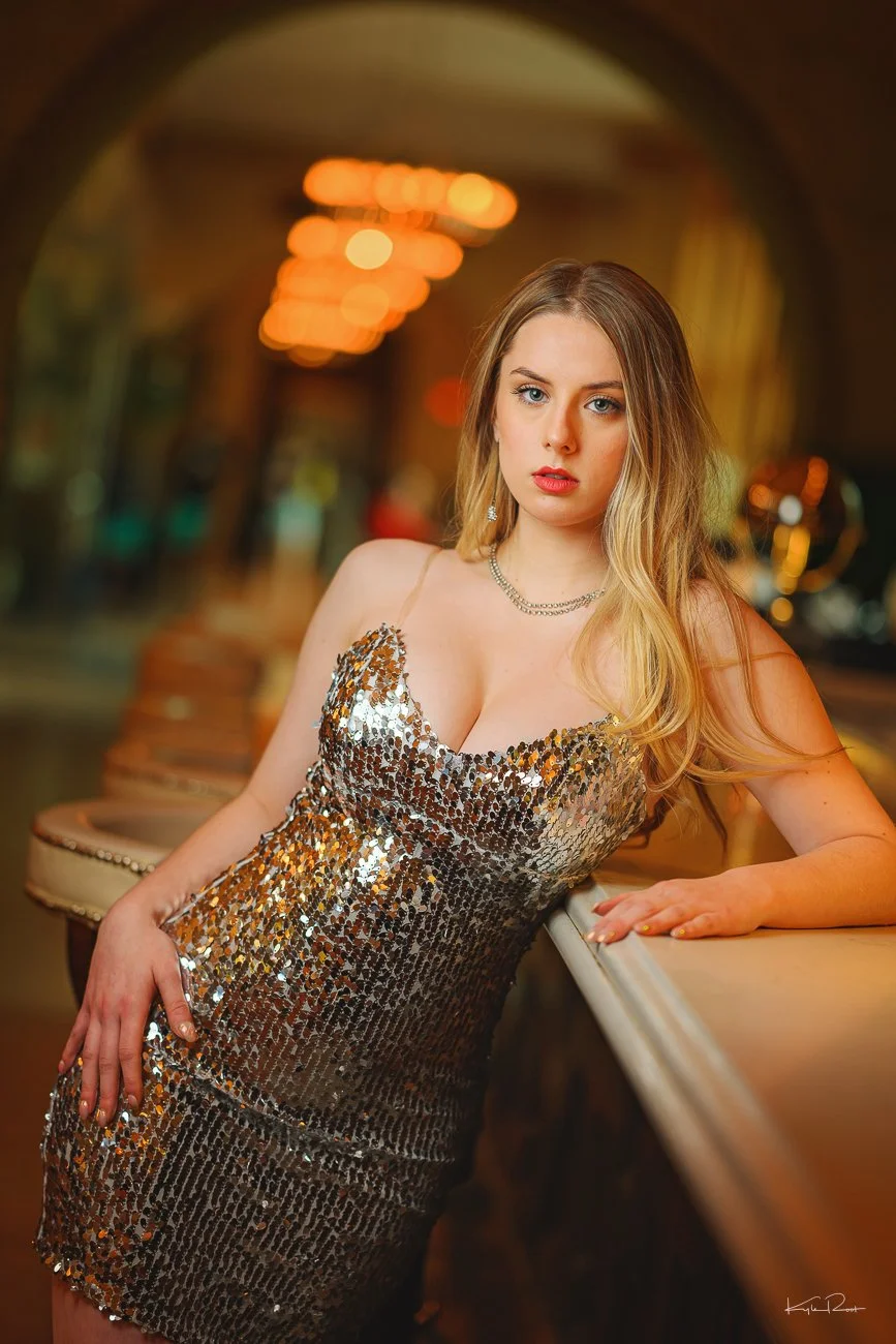 A young woman with long blonde hair wearing a sleeveless, silver sequined dress, standing in a dimly lit indoor setting with warm lighting and blurred background lights.
