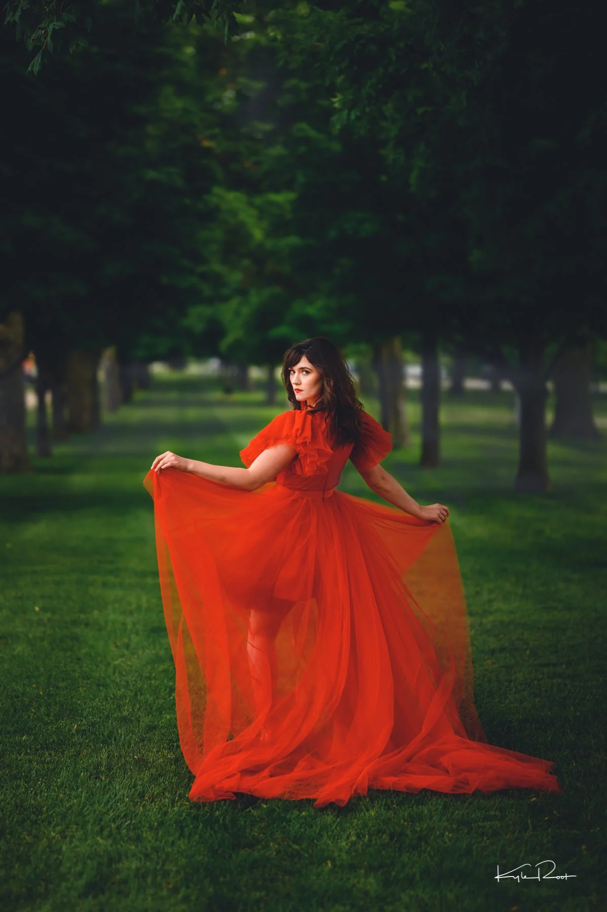 Woman in a flowing red dress standing on grass in a park with trees in the background.