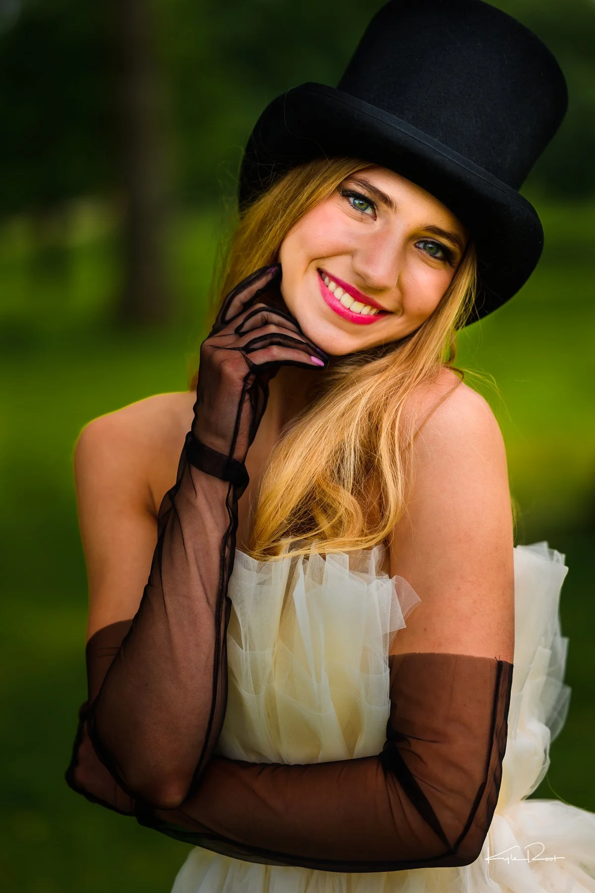 A young woman with long blonde hair, wearing a black top hat, sheer black gloves, and a white garment with ruffled fabric, smiling outdoors with greenery in the background.
