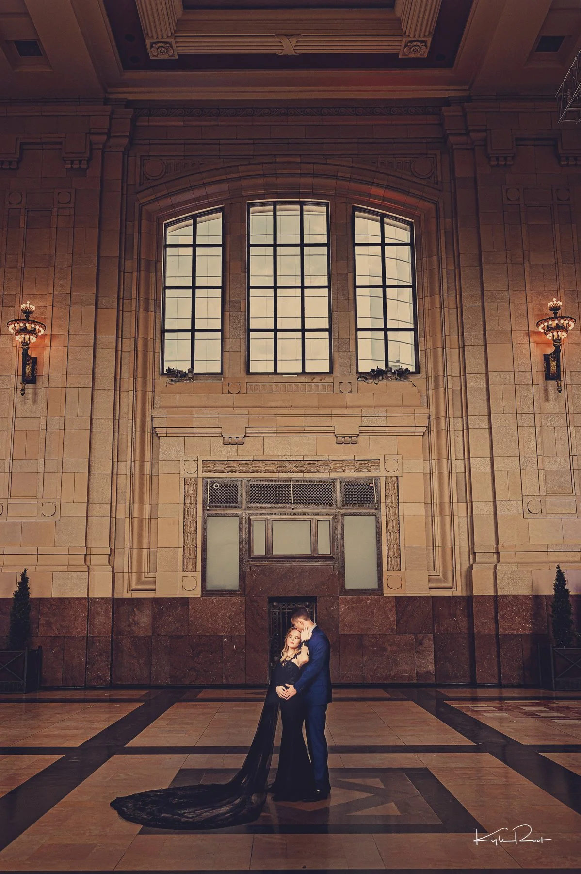 A couple dressed in formal attire standing close together in an elegant, grand hall with high ceilings, large arched windows, and ornate lighting fixtures. The woman is wearing a black gown with a long train, and the man is in a blue suit. They are e