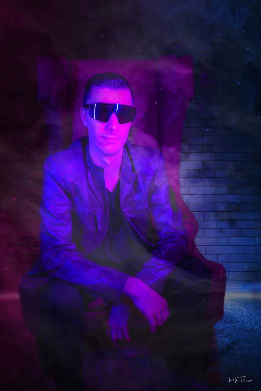 A man wearing sunglasses and a leather jacket sitting on a red chair in a dimly lit room with neon purple and blue lighting, creating a futuristic or cyberpunk atmosphere.