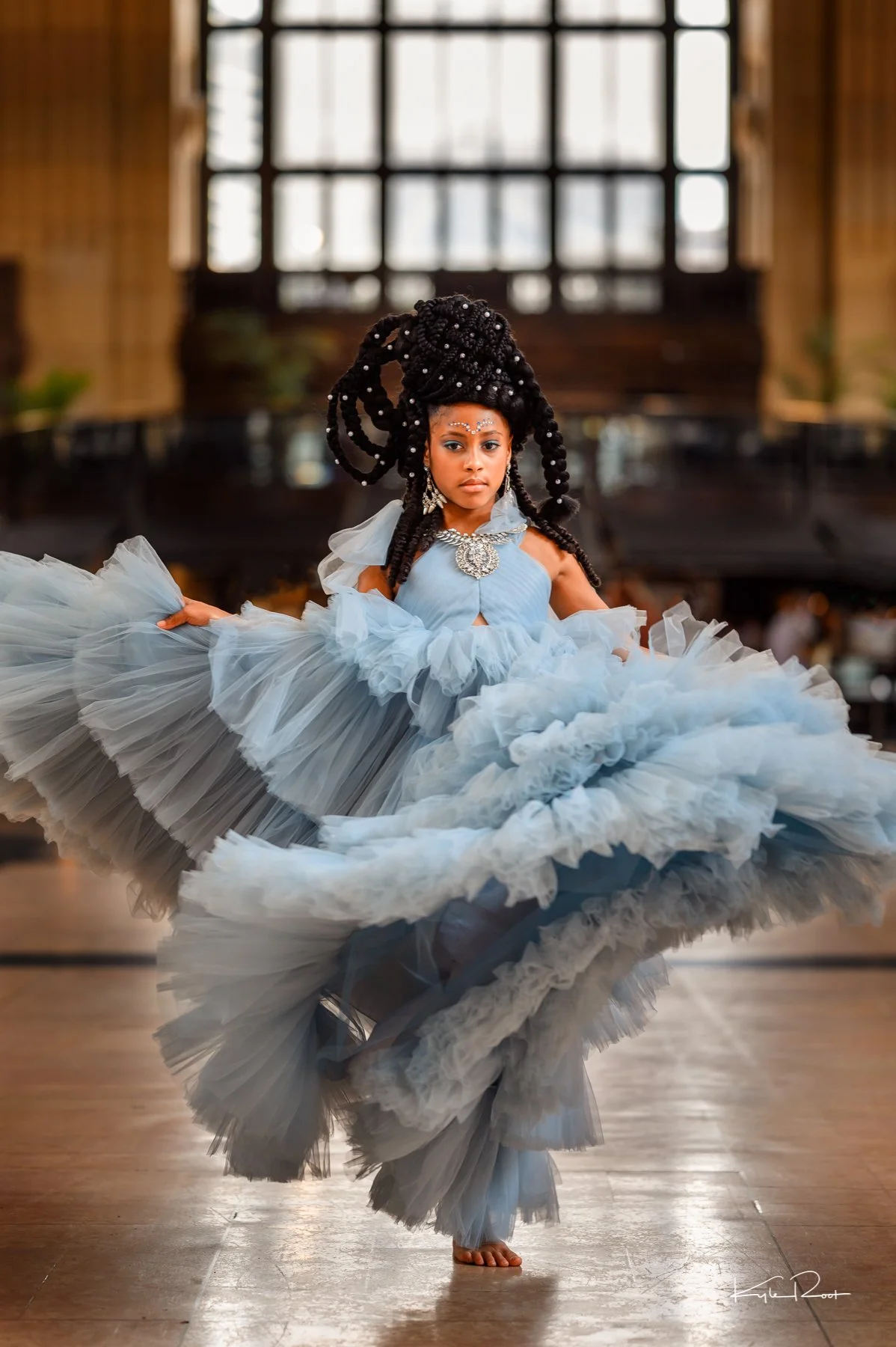 A young girl in a flowing, layered light blue dress dancing in a spacious indoor setting with large windows in the background.