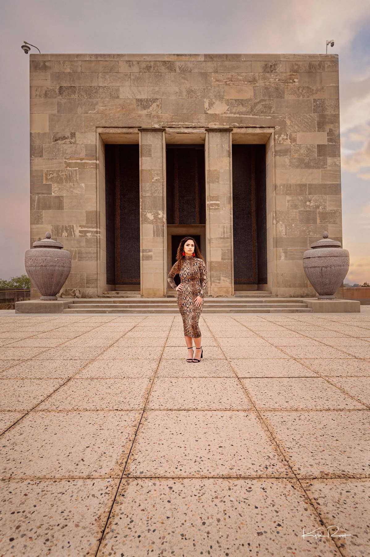A woman in a leopard print dress and high heels standing on a paved area in front of a large stone building with steps and two large urns on either side.