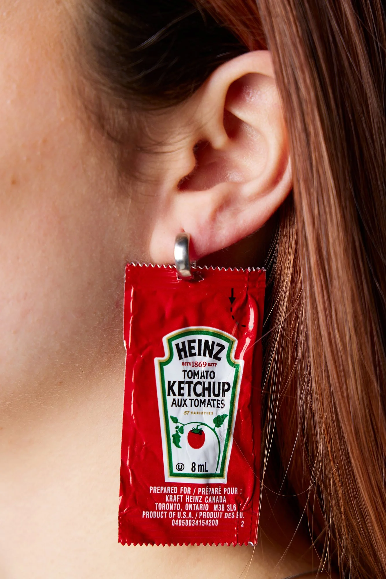 Closeup of Heinz ketchup packet hanging from small silver earring on young woman with strawberry blonde hair.