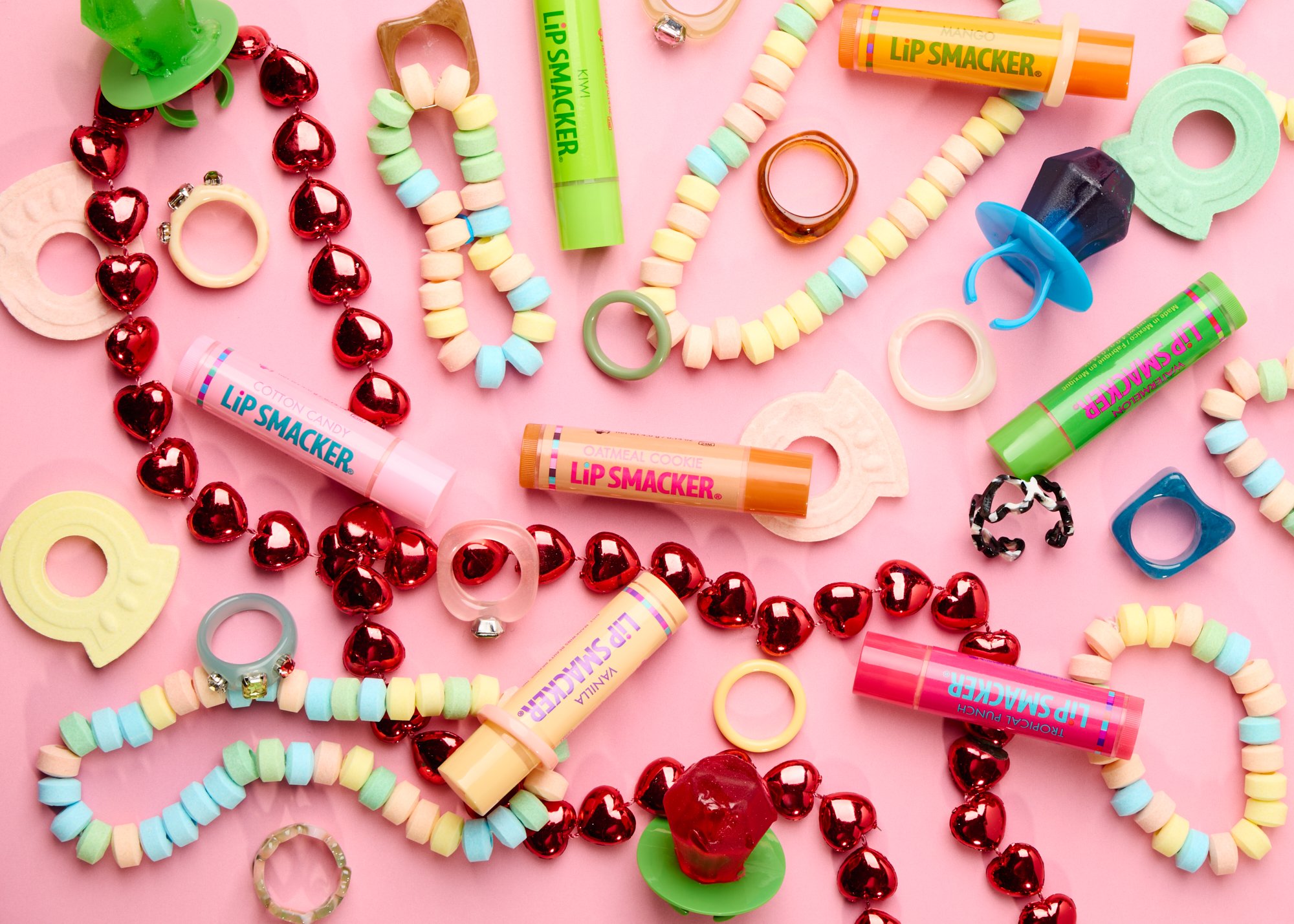Lip Smacker lip balm with ring pops, candy necklaces and bracelets, red heart beads and plastic rings scattered on pink background.