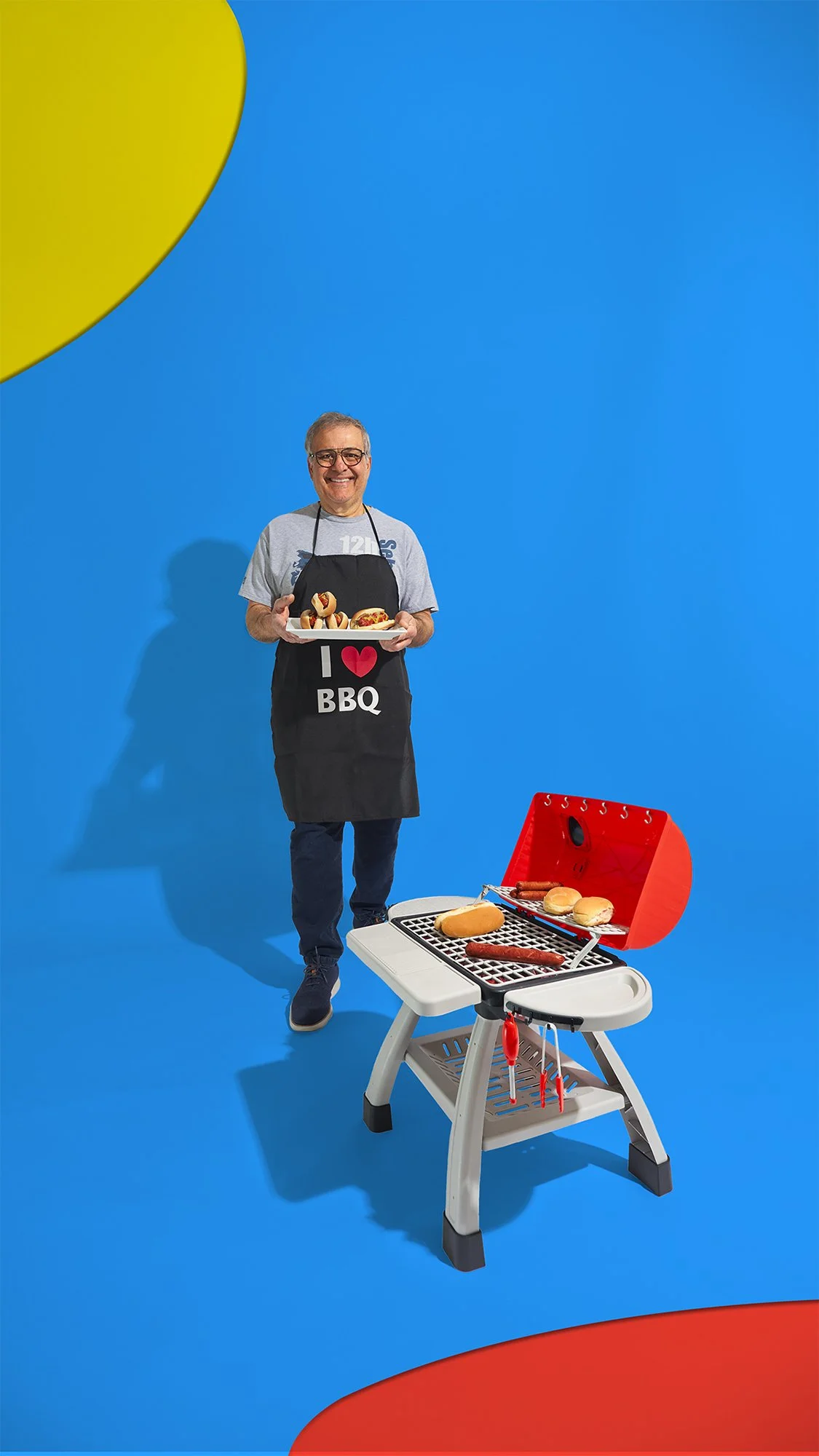 Older man wearing "I ♥ BBQ" black apron holding tray of hot dogs walking towards red and grey toy barbecue grill with food. Blue background with yellow and red abstract shapes.