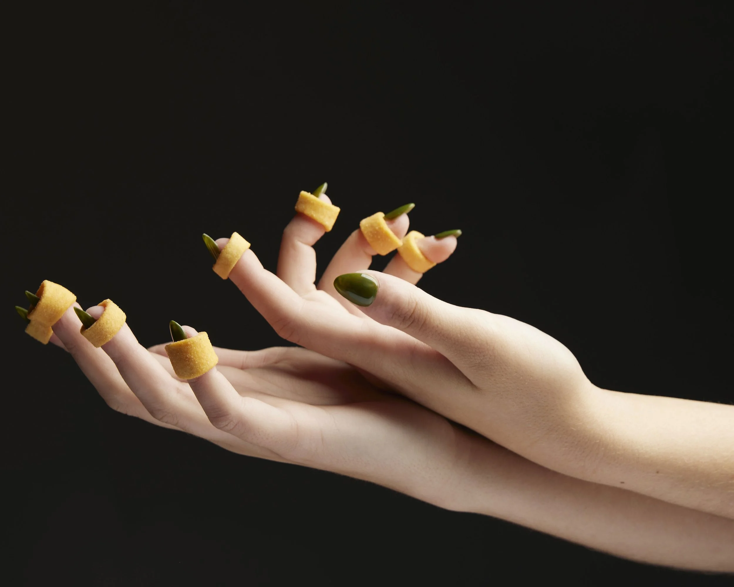 Pair of female hands with green nail polish and Ringalos on each finger. High contrast lighting on black background.