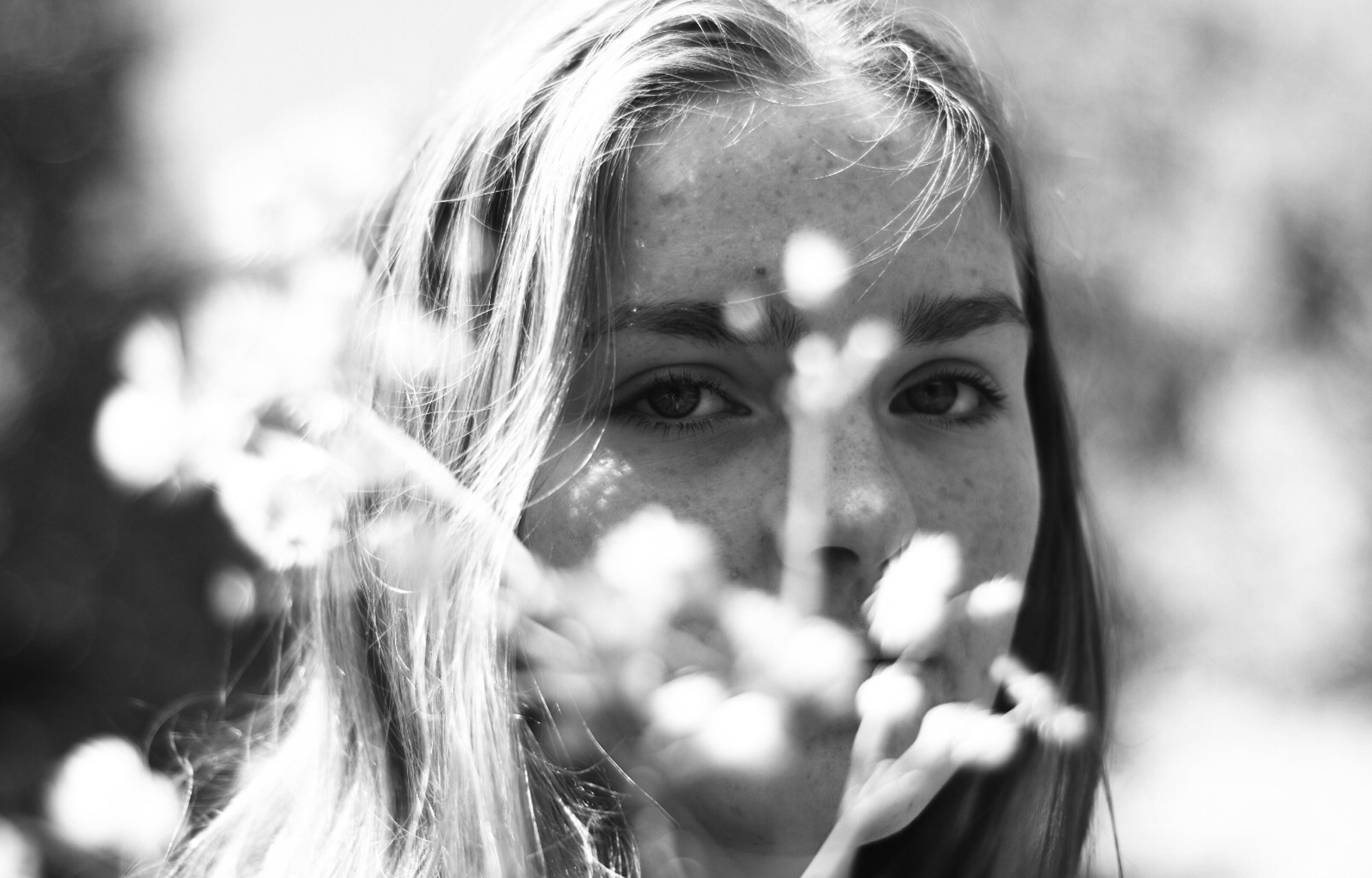 Black and white image of young white woman with blonde hair blowing in the breeze with flowers in front of her face.