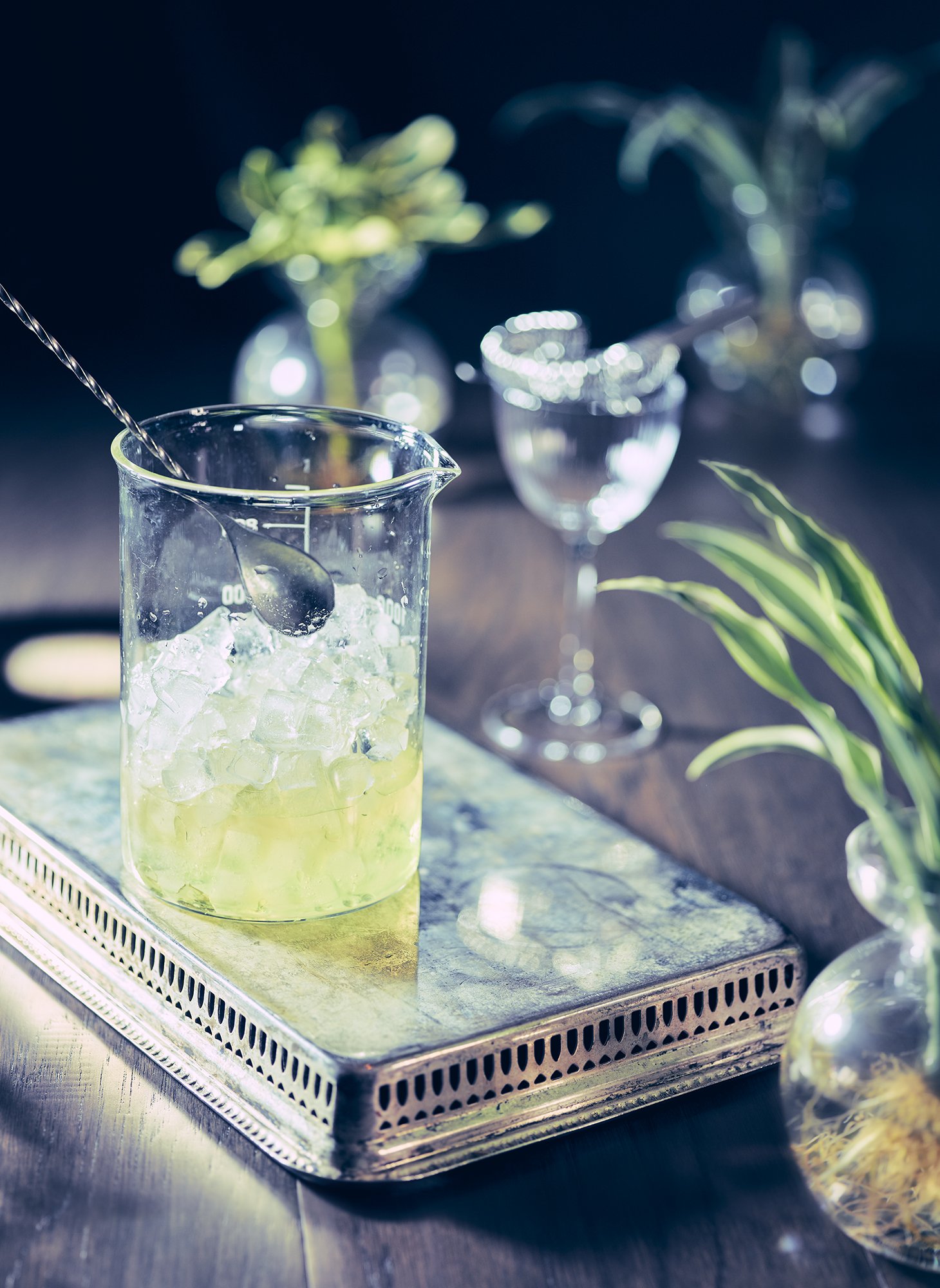 Cocktail featuring pale green fennel cocktail in laboratory beaker with crushed ice, on silver tray with glassware and herbs.