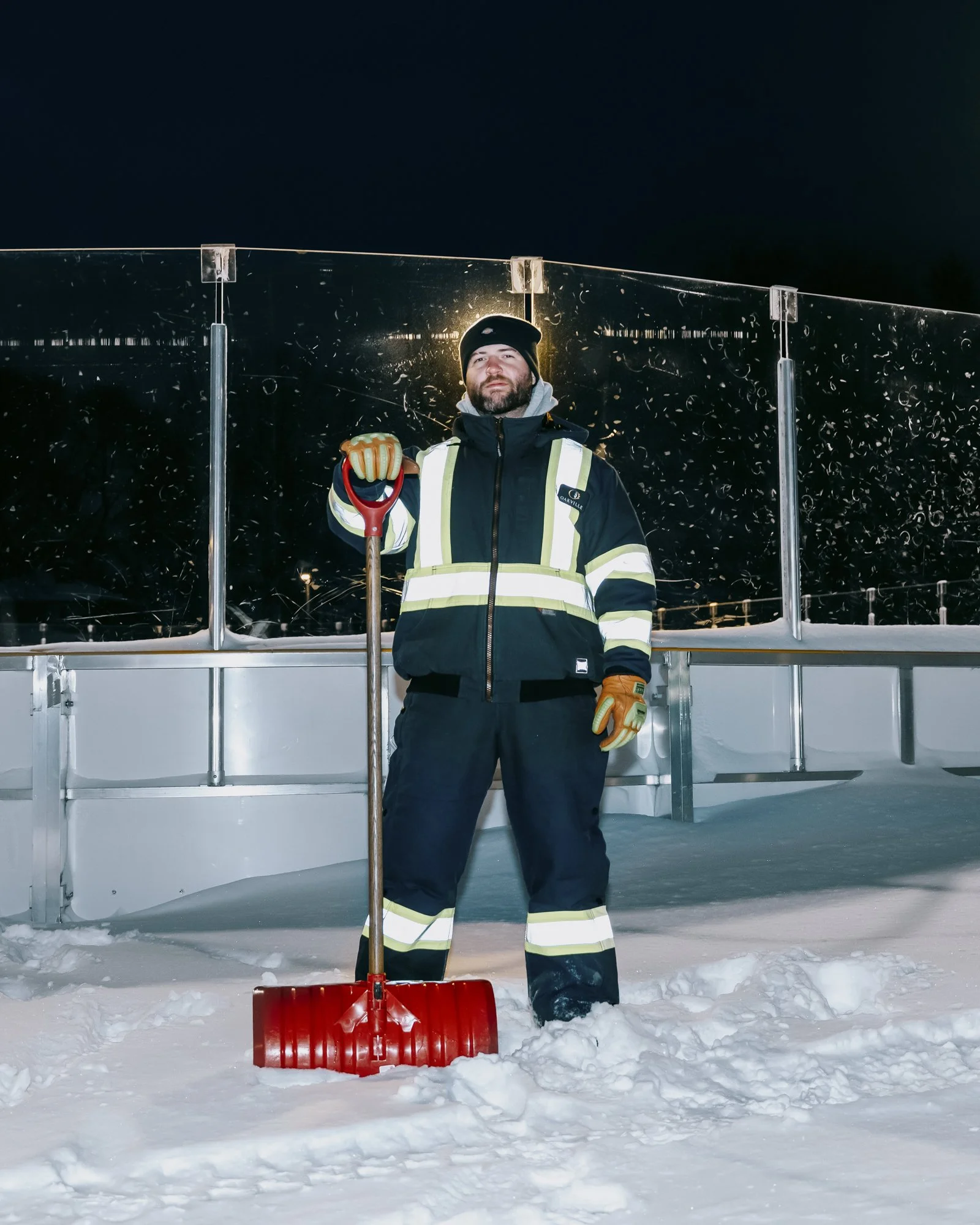 Man in hi-vis gear holding red snow shovel, standing in front of outdoor ice rink on a winter night.