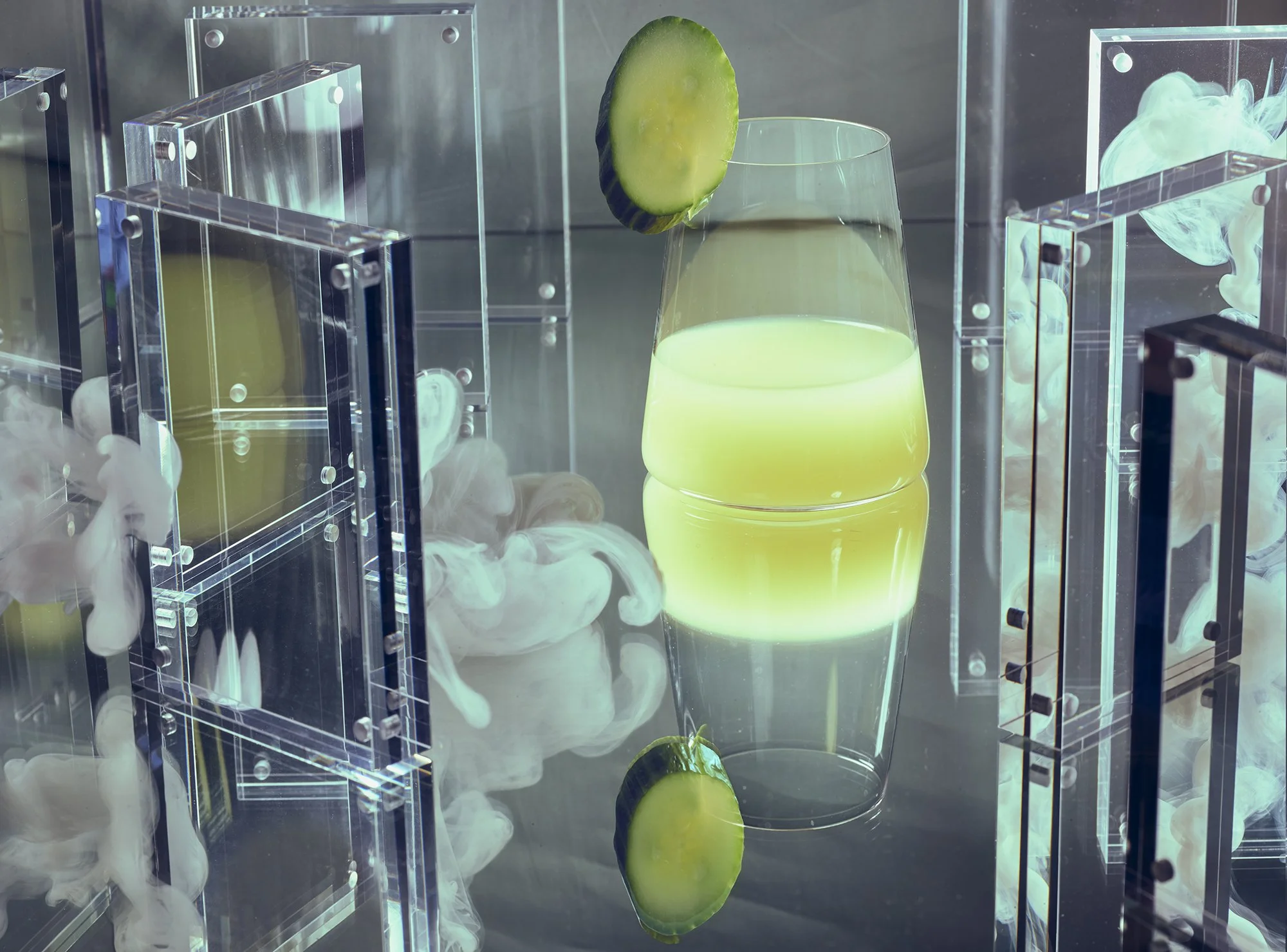 Pale yellow cocktail with cucumber surrounded by acrylic blocks and clouds.