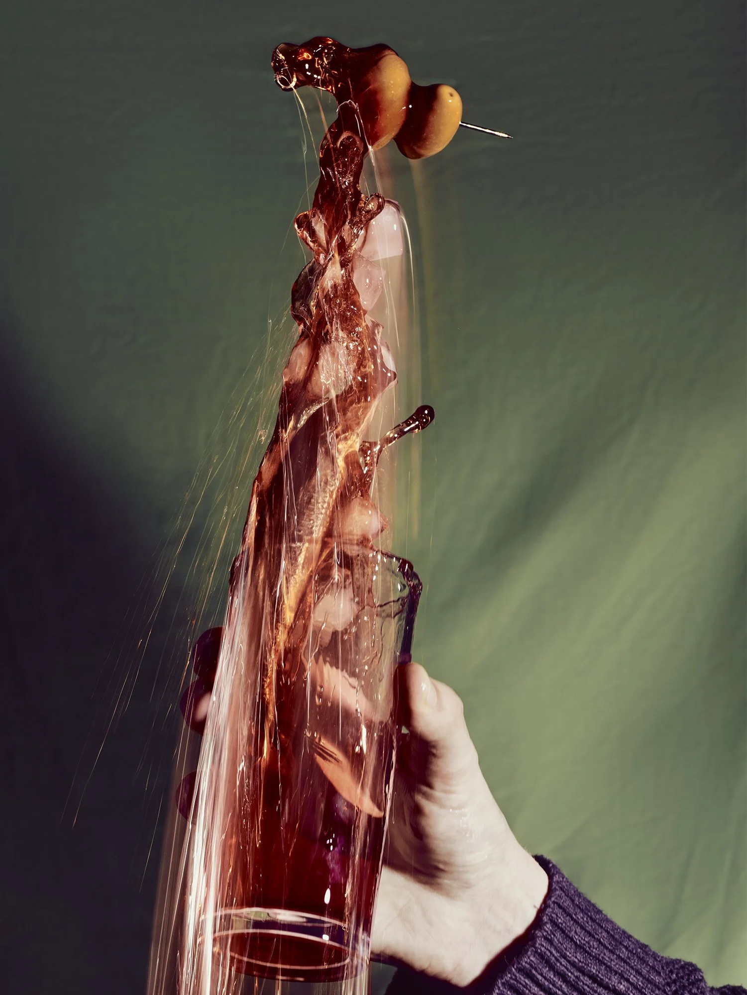 Martini photograph showing liquid being poured, olive garnish, with dragged shutter and strobe lighting. Motion blur and prismatic reflections against dark background.