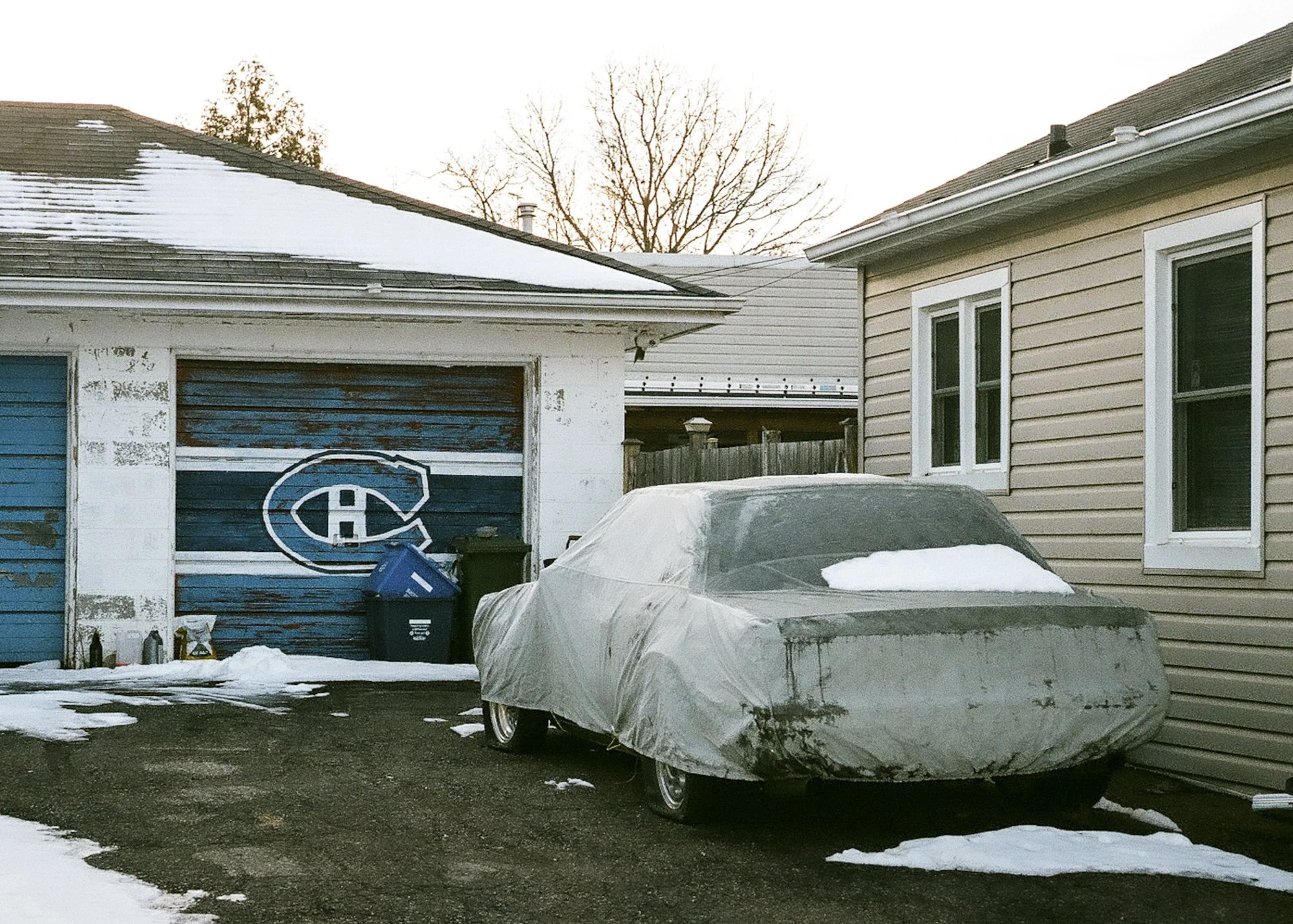 Car covered by tarp sitting in driveway, partially covered in snow in front of garage with a blue Montreal Canadiens logo.