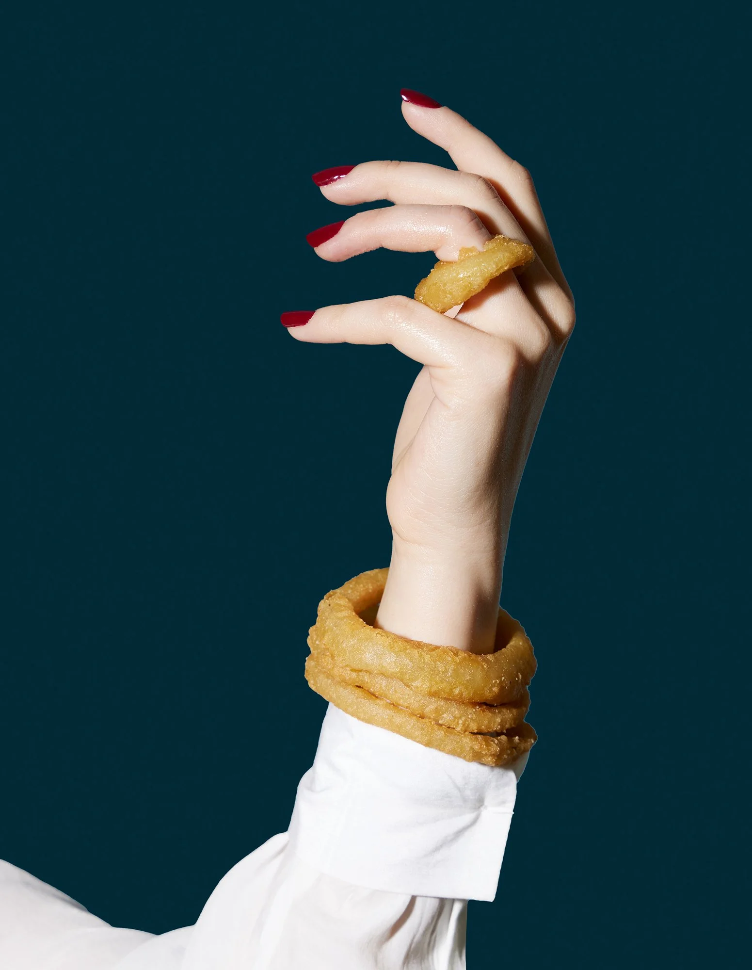 Woman with red painted nails wearing white blouse holding hand upward with three onion rings on wrist as bracelets and one on ring finger against navy blue backdrop.