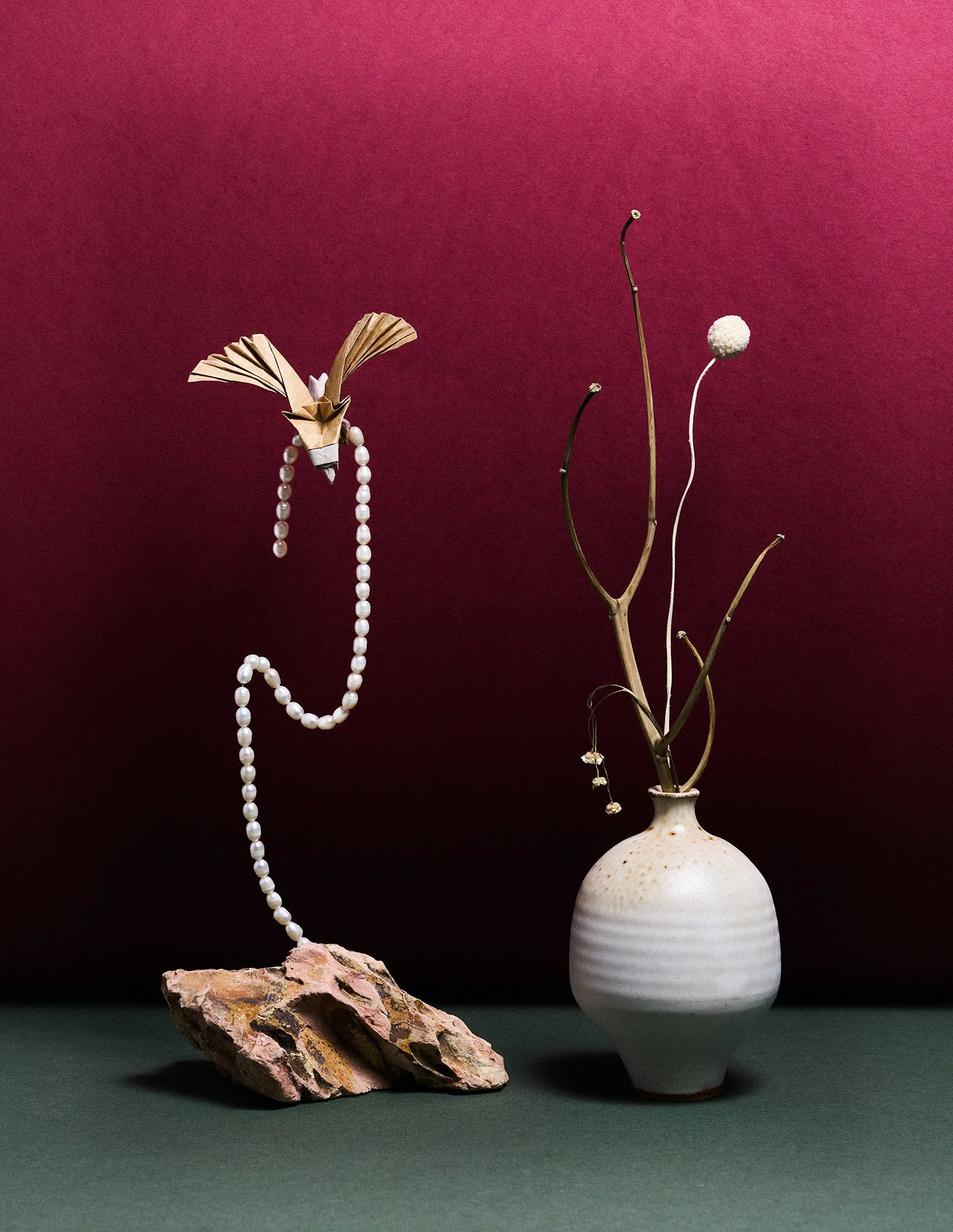 Still-life with floating string of pearls, origami bird, ceramic vase with dried branches on deep magenta ombre background.