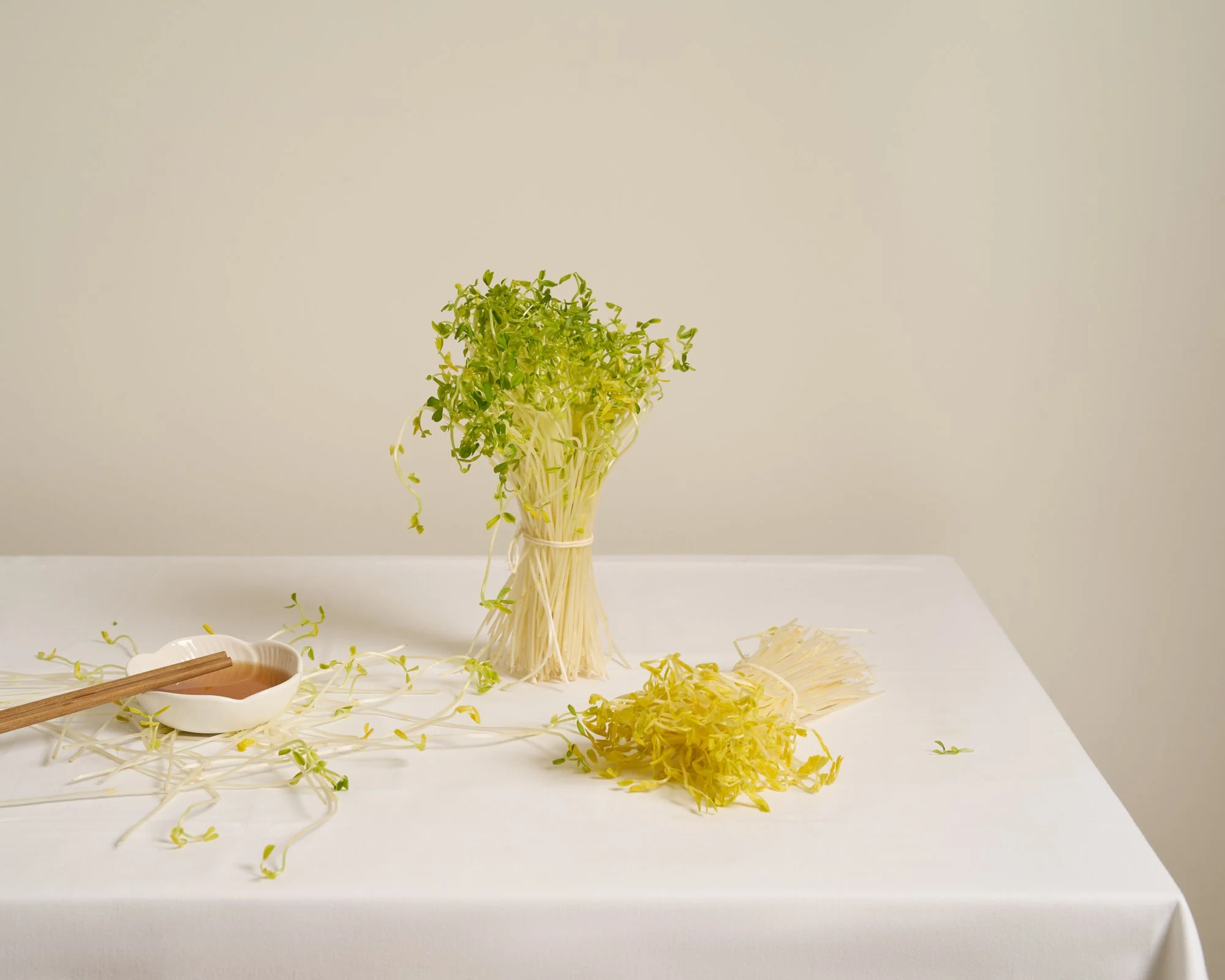 A white tablecloth-covered table with a bundle of green sprouts in the center, and shredded sprouts to the right. A small white dish with a wooden spoon containing a brown wine is also on the table.