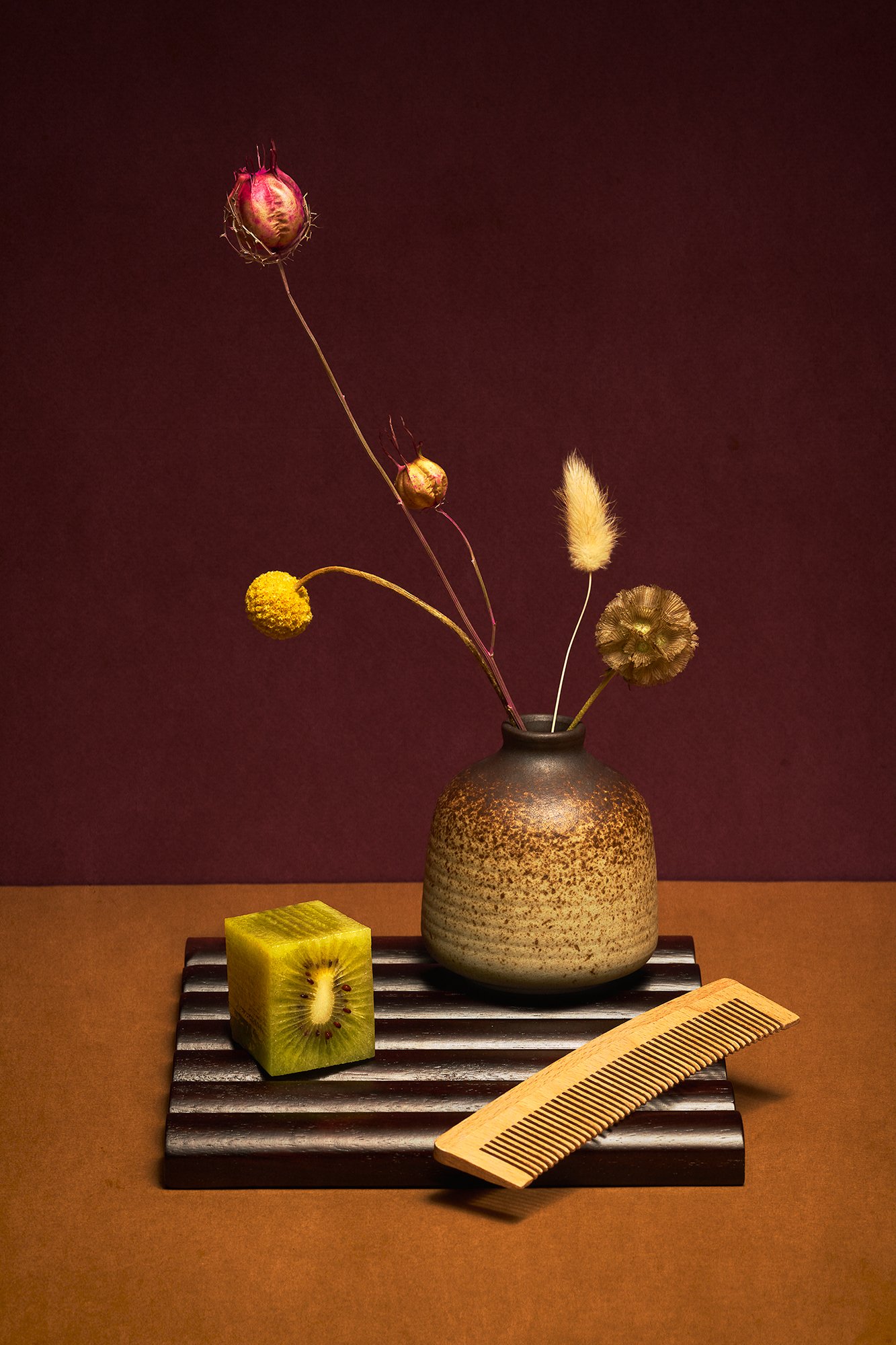 Still-life with brown ceramic vase holding dried stems on dark ridged tray with cube-cut kiwi and wooden comb, arranged on deep burgundy background.
