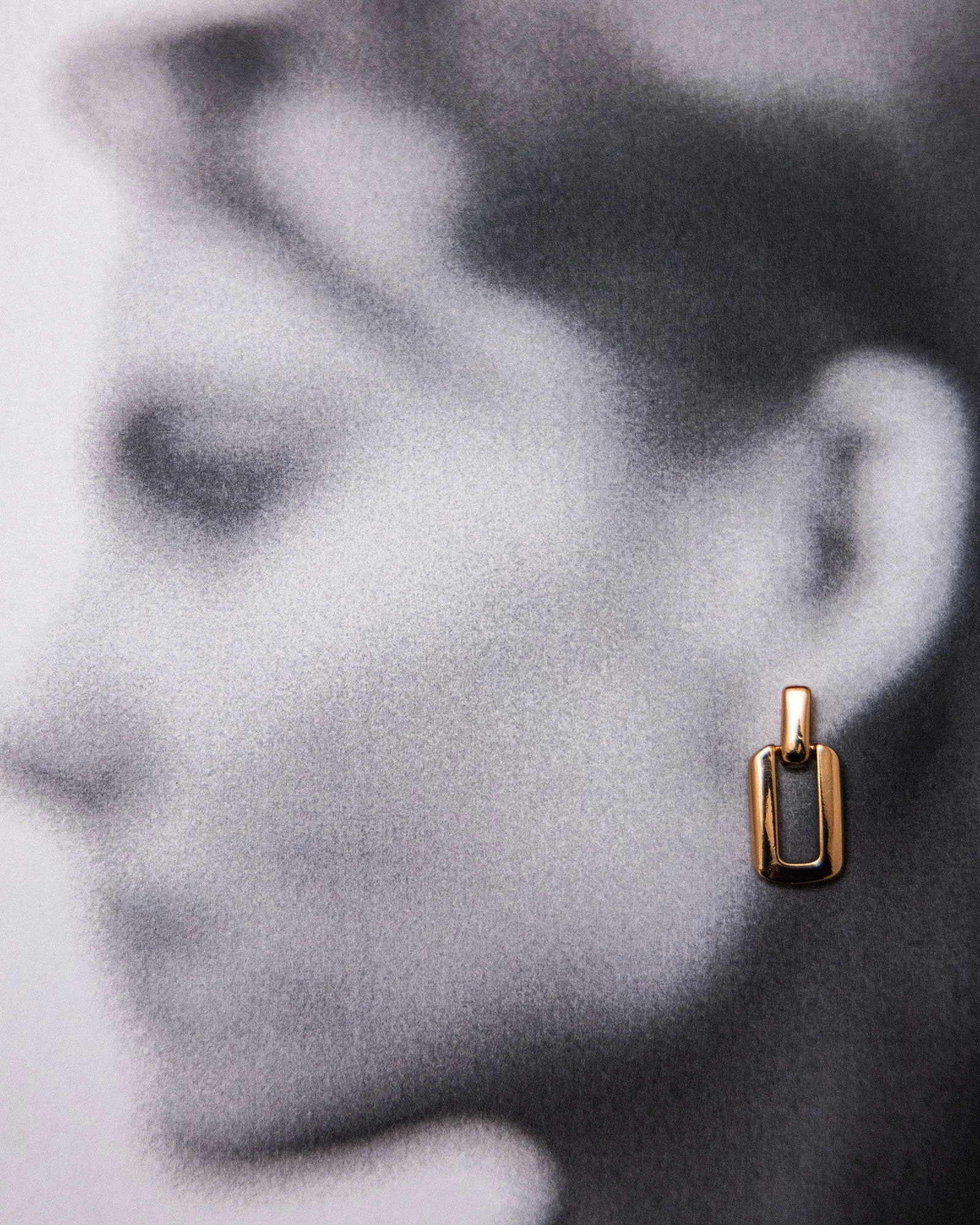 Blurred, close-up, black-and-white profile of a woman with a gold rectangular earring on the ear.