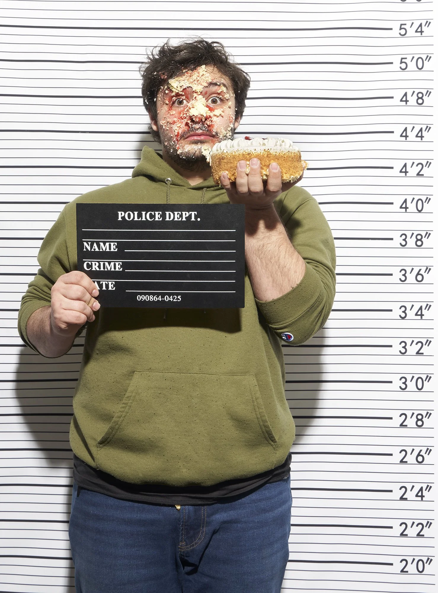 Man with face covered in cake wearing green hoodie and blue jeans. Height measurement chart in background, holding police department sign and cake.