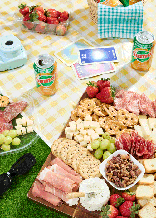 Picnic scene with charcuterie board, meats, cheese and fruit, strawberries, grapes, Sumol cans and cards on yellow checkered picnic blanket, hand reaches in, grabs pretzel and dips into cheese.