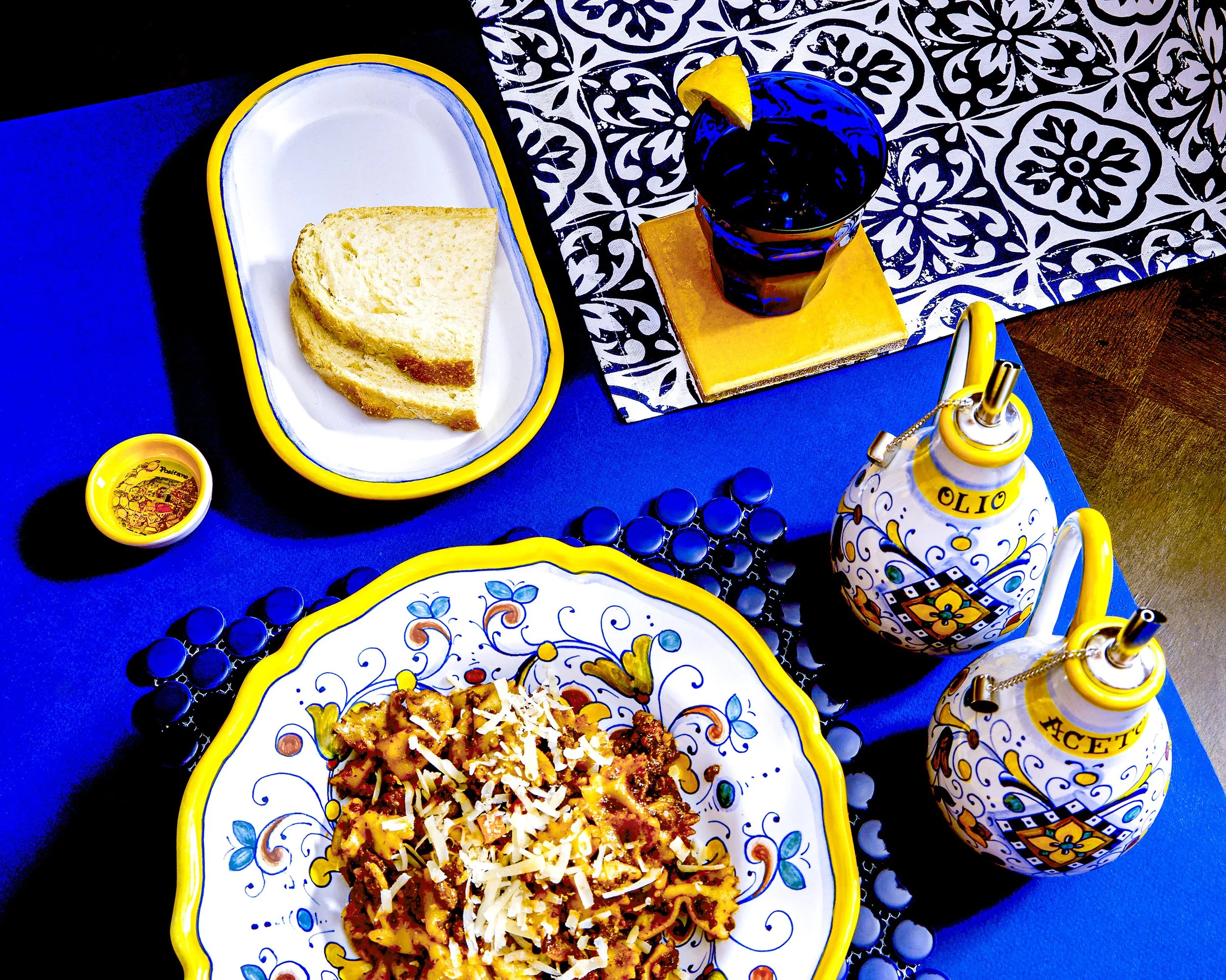 Bowl of pasta, yellow drink coaster with blue glass and slice of lemon. Bread on white side plate, oil and vinegar dispensers, blue placemat on brown wood table, high contrast lighting.