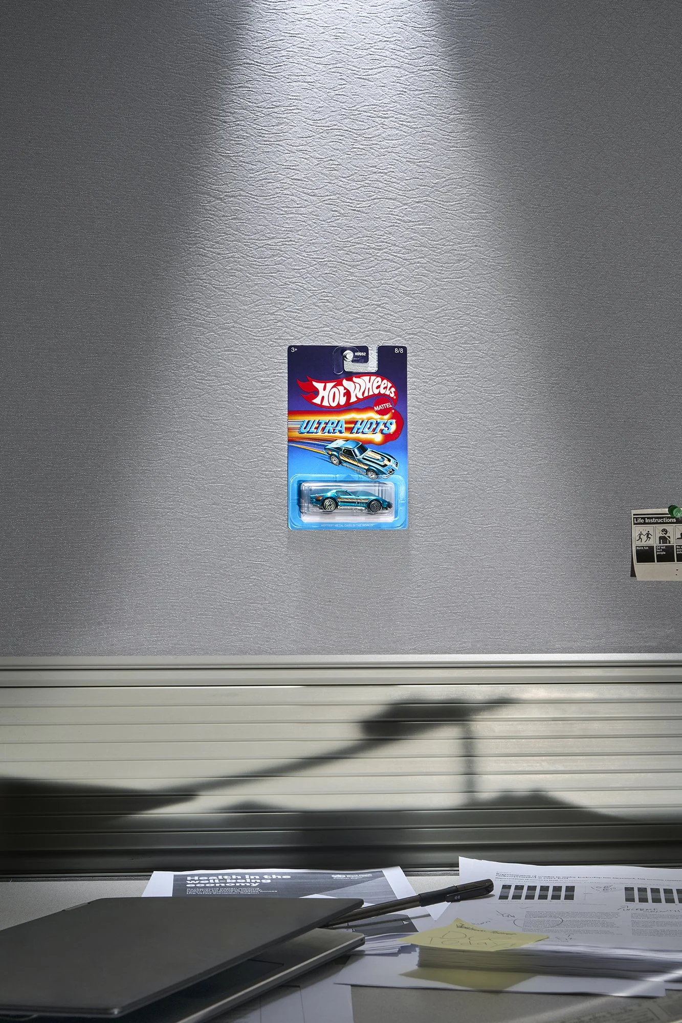 Hot Wheels toy car in package mounted to grey cubical wall above desk. Desk contains laptop, papers, black pen. large shadow cast across wall displaying toy car driving on ramp.